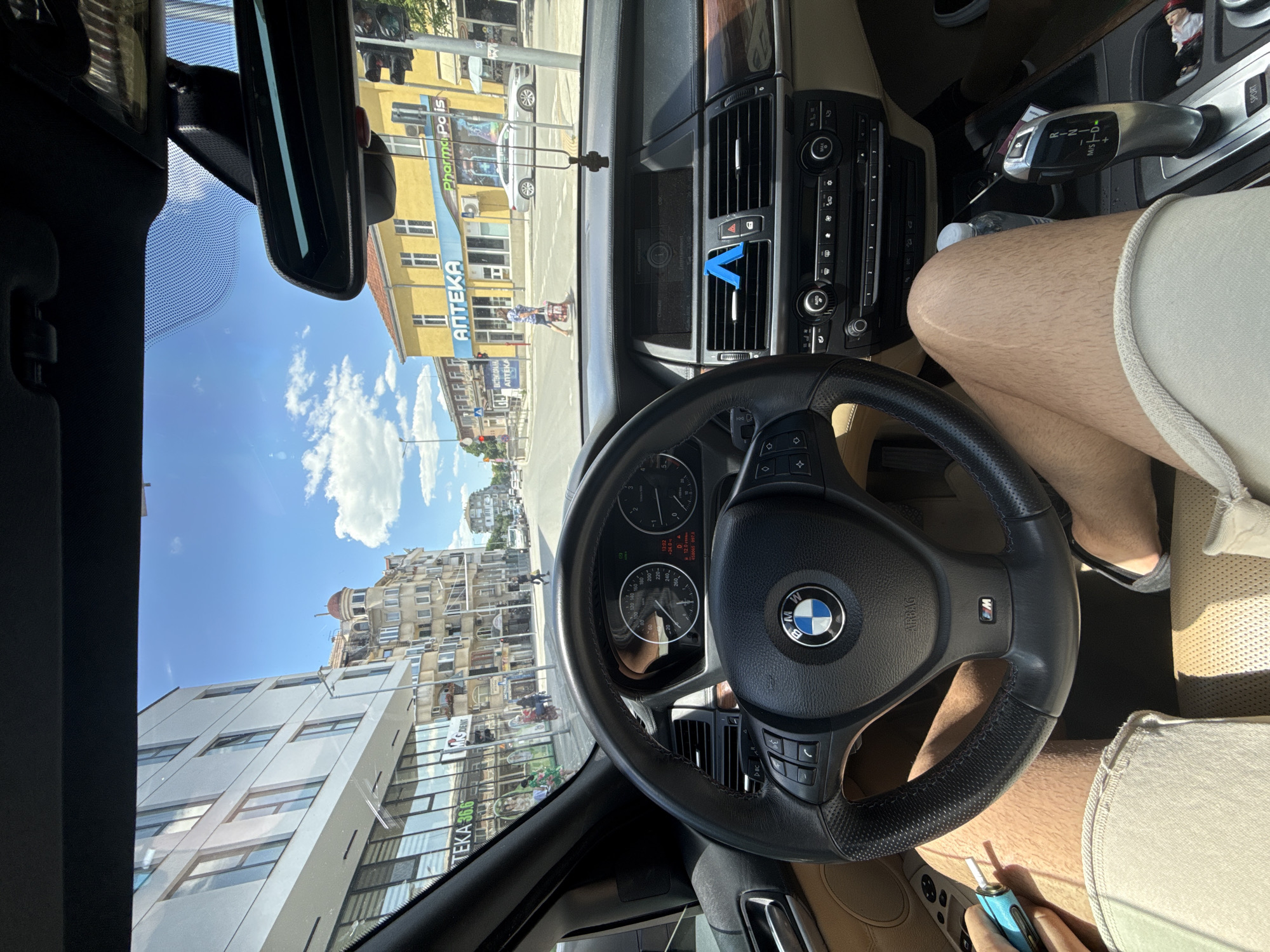 Here is a detailed description of the image:
**Scene Overview:** The image is taken from the driver's seat of a BMW, with a view through the windshield of a street scene. The city street, captured through the windshield, includes buildings, sky, and signs, giving context to the location.
**Location:** The scene is located in Plovdiv, Bulgaria. The most notable landmarks that confirm this is an "Аптека" (Pharmacy) sign in Cyrillic on a building, and another sign "TEKA 36.6" which is a pharmaceutical brand.
**Interior Details:** The vehicle's dashboard, steering wheel, and center console are visible. The steering wheel features the BMW logo, and the instrument cluster is also visible. A "W" logo is on the wheel. Air vents and parts of the car's control panel are in view. A blue device with a white tip is in the driver's hand. The driver is wearing neutral-colored shorts. The car's interior is tan.
**Exterior Environment:** Several multi-story buildings, including a white building with a glass facade and a yellow building are visible. The sky is blue with scattered clouds.
**Objects:** Besides the buildings and car components mentioned, there are also street lights.