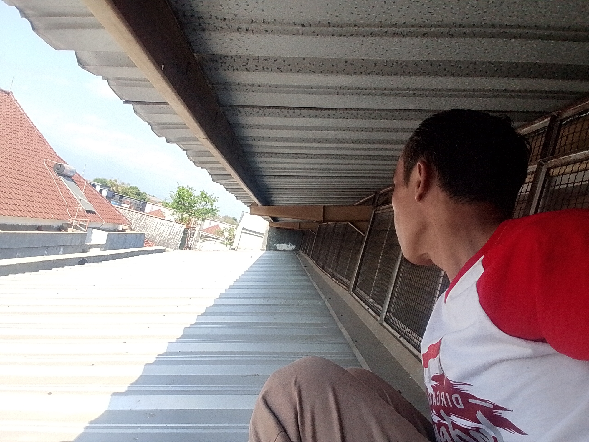 A man with short, dark hair is pictured from the back, looking to his right. He is wearing a white t-shirt with red sleeves and a red graphic design on the front. He is also wearing brown pants. The man is sitting on a corrugated metal roof. The roof is white and catches the sunlight, creating a bright, high-contrast scene. Beyond the roof, a tiled roof of a house is visible, along with some trees and a bright blue sky. The image appears to have been taken outdoors, possibly on a sunny day. Based on the context, this image may have been taken in Pasuruan, Indonesia.