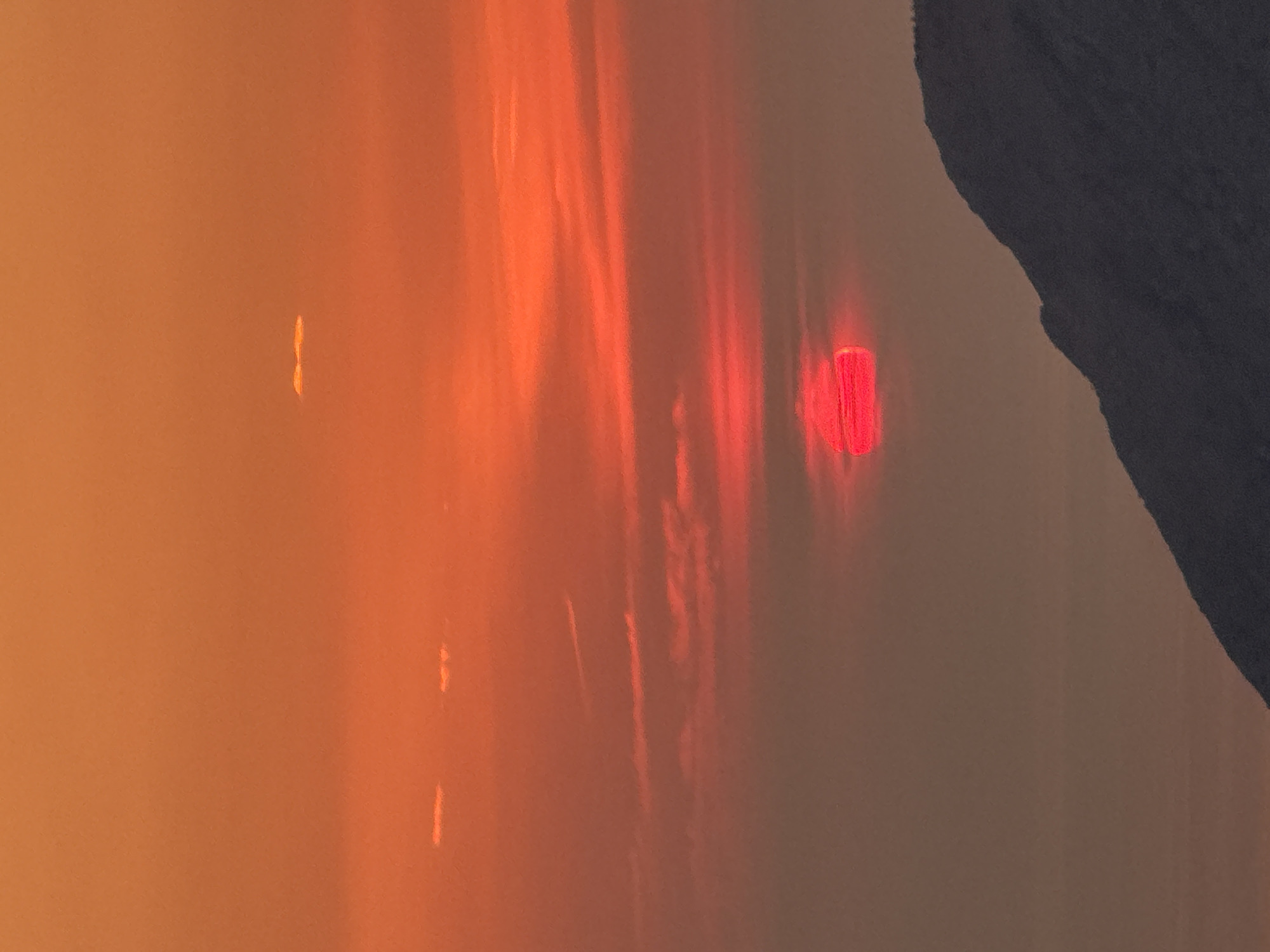 The image depicts a sunset over a mountainous landscape in Gluhi Do, Montenegro. The sky is filled with vibrant shades of orange and red, with thin, wispy clouds scattered across the expanse. The sun, a brilliant red disc, is partially obscured by a layer of haze, creating a soft, diffused glow. In the foreground, the dark silhouette of a mountain range rises against the colorful sky, its rugged contours sharply defined. The scene evokes a sense of peacefulness and natural beauty.