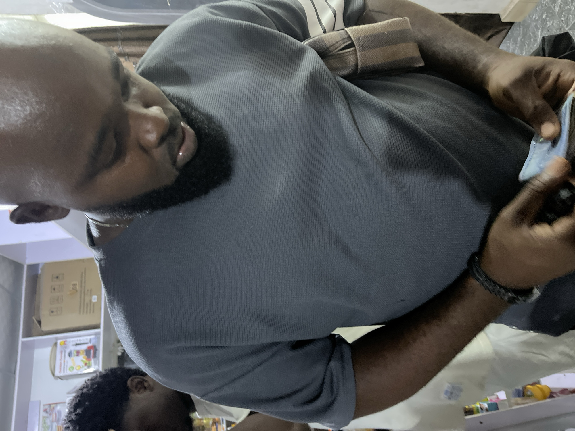 A man is seen in the foreground wearing a gray shirt with a brown and beige striped strap across his shoulder. He appears to be counting or handling what looks like money. He is dark-skinned, with a beard and a bracelet on his wrist.
In the background, there is a shelf filled with cardboard boxes, and other smaller items, suggesting a possible storage or retail environment. It is not possible to identify the specific location in Dushepe, Nigeria from the details provided in the image.