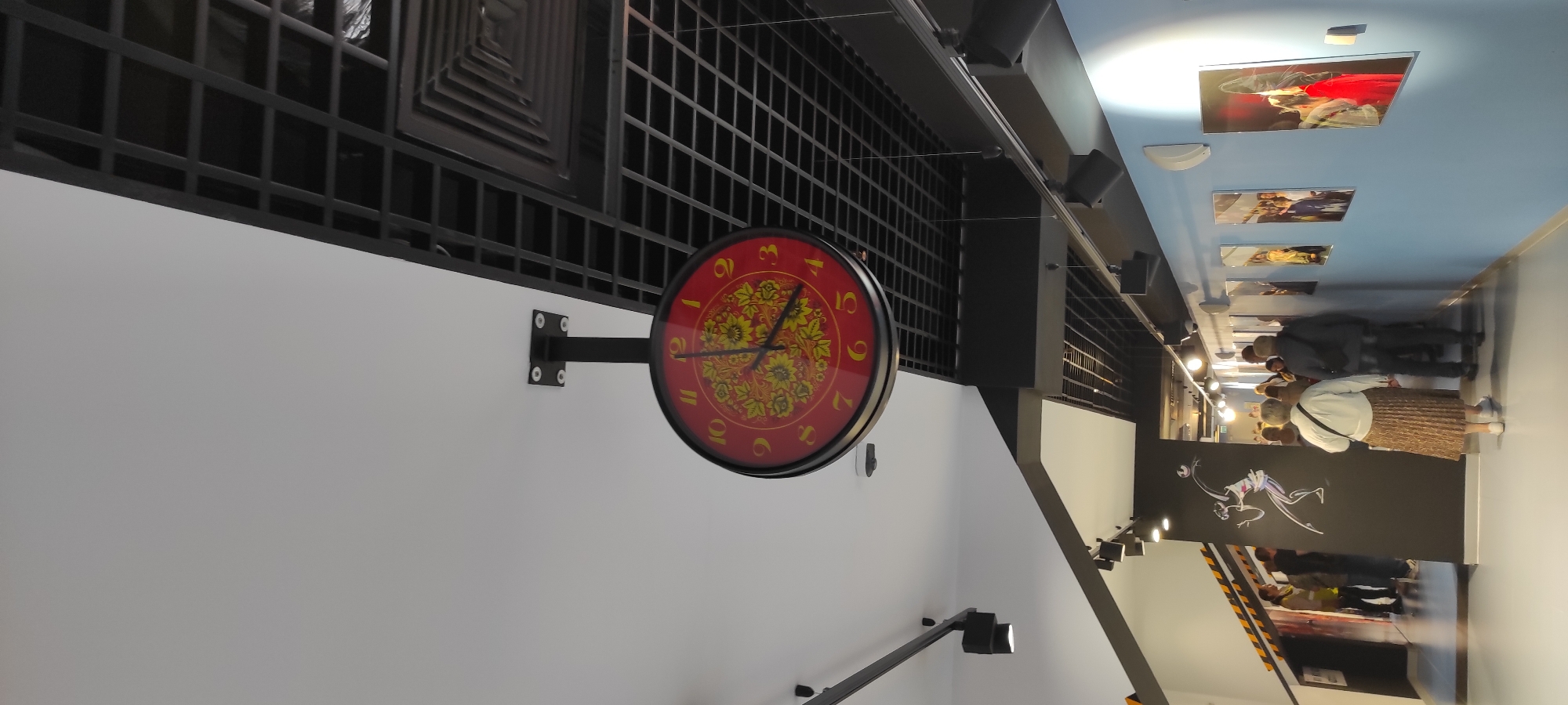 The image shows an interior scene in a modern building, likely an exhibition hall or museum, with a high ceiling and a black grid-like structure above. A prominent double-sided red clock with yellow floral patterns is suspended from the ceiling in the center of the frame. The clock face displays Arabic numerals. Below the clock, a group of people are walking down a brightly lit corridor lined with framed photographs on a light blue wall. The corridor appears to lead into another area where more people are visible. There is a graphic of a stylized human figure on a black wall segment to the left. Lighting fixtures are visible along the ceiling and walls, and a ventilation system is integrated into the black grid structure. The overall impression is one of a contemporary public space with an artistic or cultural focus.