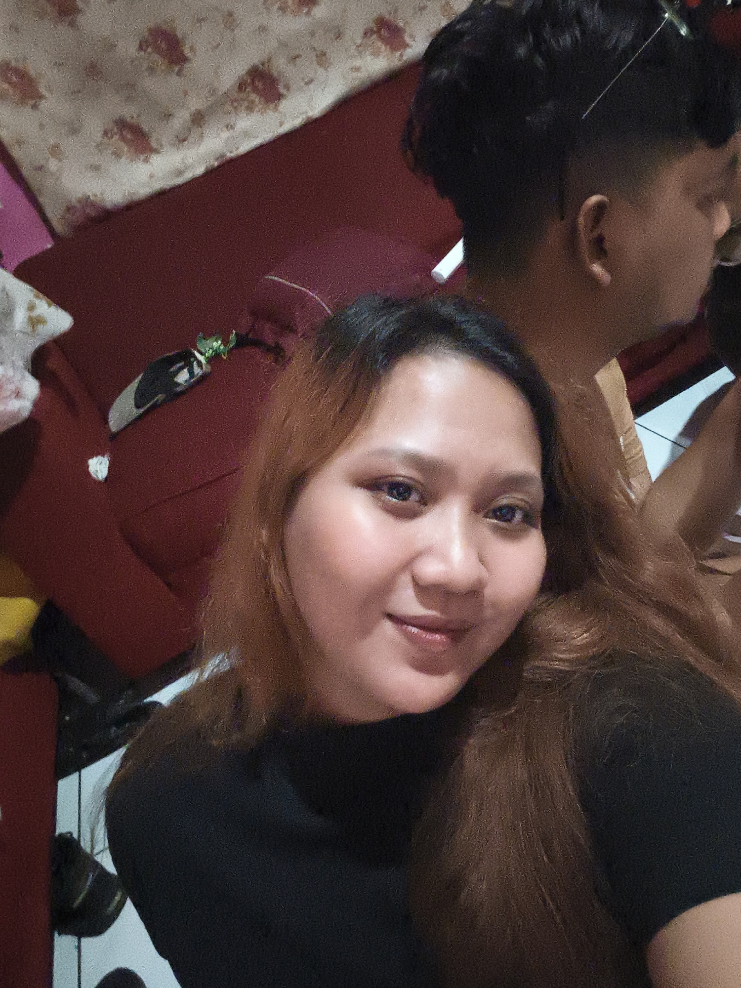 The image depicts a selfie-style photograph featuring two individuals in an indoor setting. The foreground is dominated by a woman with long, reddish-brown hair, wearing a black top. She is smiling and looking directly at the camera. Her facial features include dark eyes and light skin. In the background, partially visible, is a man with dark, short, styled hair. He is turned away from the camera, with his head angled to his right. He appears to be wearing a light-colored garment.
The environment suggests a residential space. Behind the woman, a dark red sofa is visible, with a patterned fabric (possibly a blanket or upholstery) draped over the back of it. The pattern appears to consist of floral or abstract shapes in muted colors. To the left of the sofa, a pink surface is partially visible. The floor in the lower portion of the image is tiled with white and black squares. There are also some dark objects on the floor that resemble shoes. The overall lighting is warm, suggesting artificial indoor illumination. The photograph does not provide explicit details indicating a specific location within Depok, Indonesia, but the domestic setting is evident.