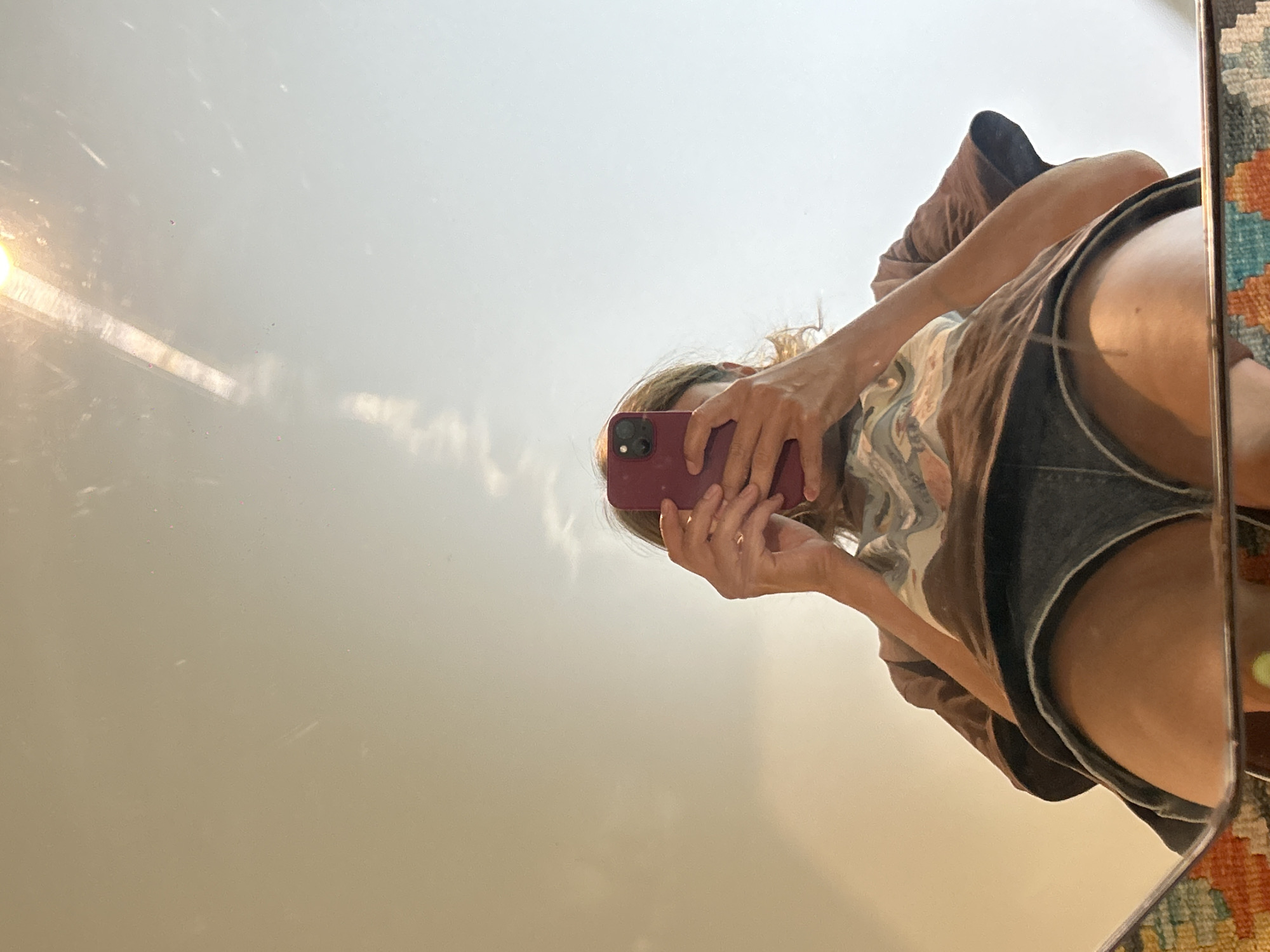 A person takes a selfie in a mirror. The person is wearing shorts and a t-shirt. A smartphone is held in their hands. The background shows an outdoor scene with hazy light, likely due to smoke from the forest fires in North Kuta, Indonesia.