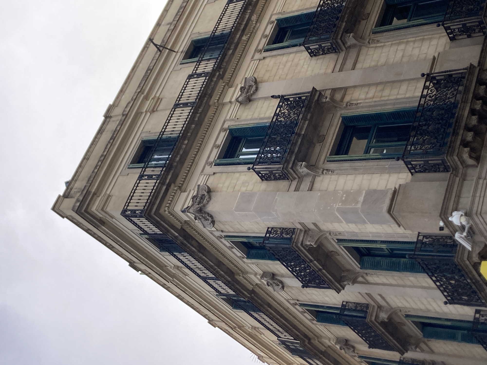 This media file captures a low-angle view of an ornate building facade, likely in Barcelona, Spain, given the architectural style. The building appears to be old, with a pale stone exterior and decorative elements. Multiple stories are visible, each featuring rows of windows with dark, intricate wrought-iron balconies. The windows themselves have teal-colored shutters. Some of the balconies have ornate metalwork, while others are more simply designed with vertical bars. Small, sculpted decorative figures, possibly gargoyles or architectural embellishments, are visible between some of the windows.
The sky is overcast and appears to be a pale grey, suggesting it is either early morning, late afternoon, or a cloudy day. There are no people or visible activity in the frame, making it a still shot of the building's exterior. The focus is on the architectural details and the imposing structure of the building, highlighting its classical design and aged character. The perspective emphasizes the height and grandeur of the building. No text is visible in the image.