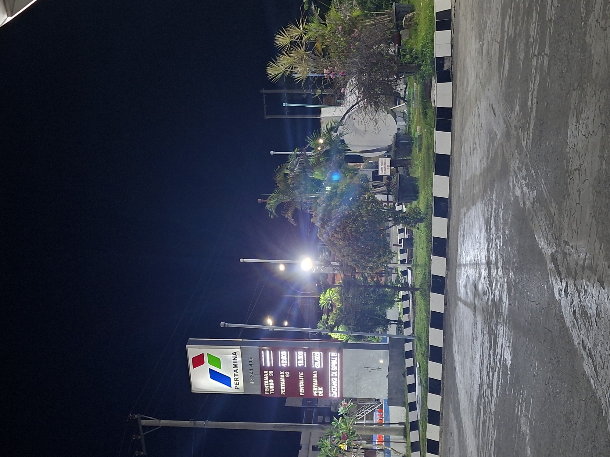 This is a night time, eye-level, exterior shot of a Pertamina gas station in Keuniree, Indonesia. The prominent feature is a tall, illuminated sign displaying the Pertamina logo and various fuel prices. The sign is lit by internal lighting, making the numbers and brand name clearly visible against the dark night sky. To the left of the sign is a utility pole with wires. In front of the sign, a paved area with a black and white striped border is visible, with grass and some potted plants. Behind the sign, palm trees and other foliage are illuminated by streetlights, creating a tropical atmosphere. A large white storage tank is visible in the background, along with more vegetation. The ground in the foreground appears wet, suggesting recent rain. There are no people visible in the scene.