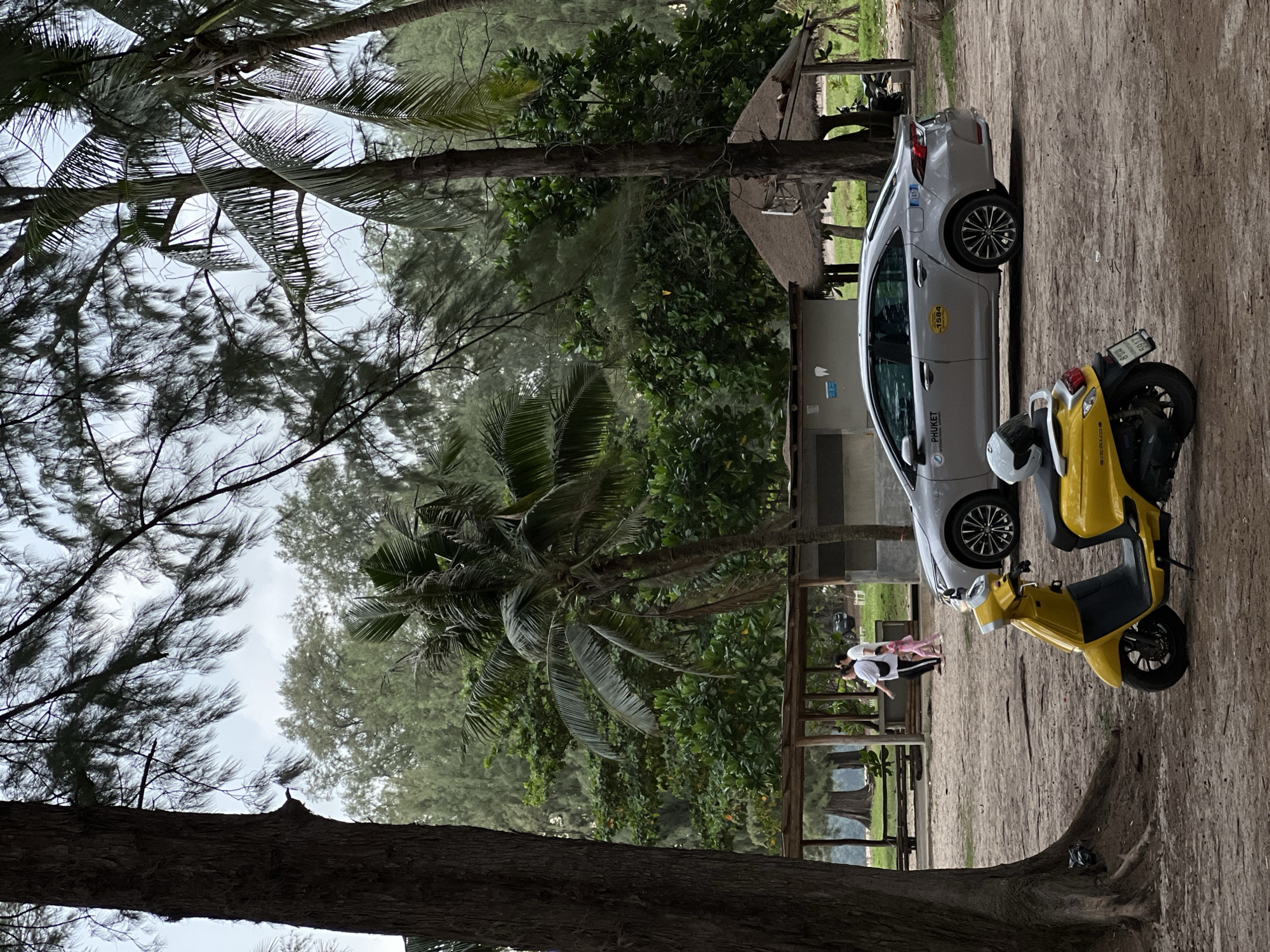 An outdoor, low-angle shot captures a tropical scene with a yellow scooter in the foreground, parked on a sandy surface. Behind the scooter, a silver sedan is parked, with a license plate that reads "1584". Two figures, a woman and a child, walk in the distance towards a structure with a thatched roof. The background is filled with lush green trees and foliage, suggesting a beachside or park environment. The sky is overcast, with branches of trees partially obscuring the view.