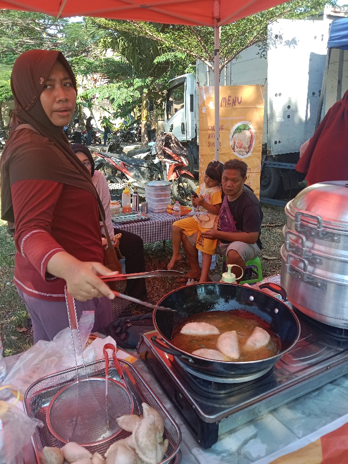 In Kebondalem, Indonesia, an outdoor food stall operates under an orange canopy during daylight hours. A woman, wearing a dark brown hijab and maroon shirt, stands in the foreground, holding red-handled tongs to fry crescent-shaped items in a black pan on a portable gas stove. To her left, a metal basket holds additional uncooked food and utensils. To her right, a stack of three silver steaming pots is visible. In the mid-ground, a man sits on a green stool, looking forward, with a young girl holding a mobile phone sitting beside him, barefoot. Behind them, another woman is partially visible seated at a checkered table displaying plastic containers and condiment bottles. The background reveals parked motorcycles, a white box truck, and a light blue/grey box truck featuring a "MENU" sign. Green foliage and trees indicate an open, outdoor setting with a dirt/grass ground.