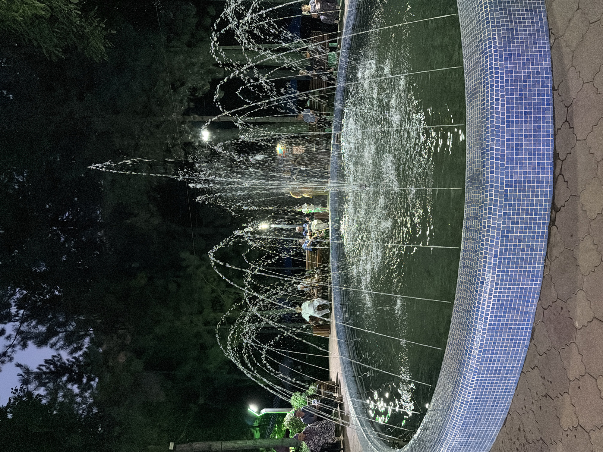 The image shows an outdoor fountain at dusk in Khumsan, Uzbekistan. The fountain is circular with a blue tiled rim and is actively spraying water in arcs. Several people are seated on benches around the fountain, observing it. The surrounding environment appears to be a park with trees and lampposts, suggesting it is a public space. The lighting indicates it is evening, with artificial lights illuminating the fountain and the area. The ground around the fountain is paved with decorative stones.