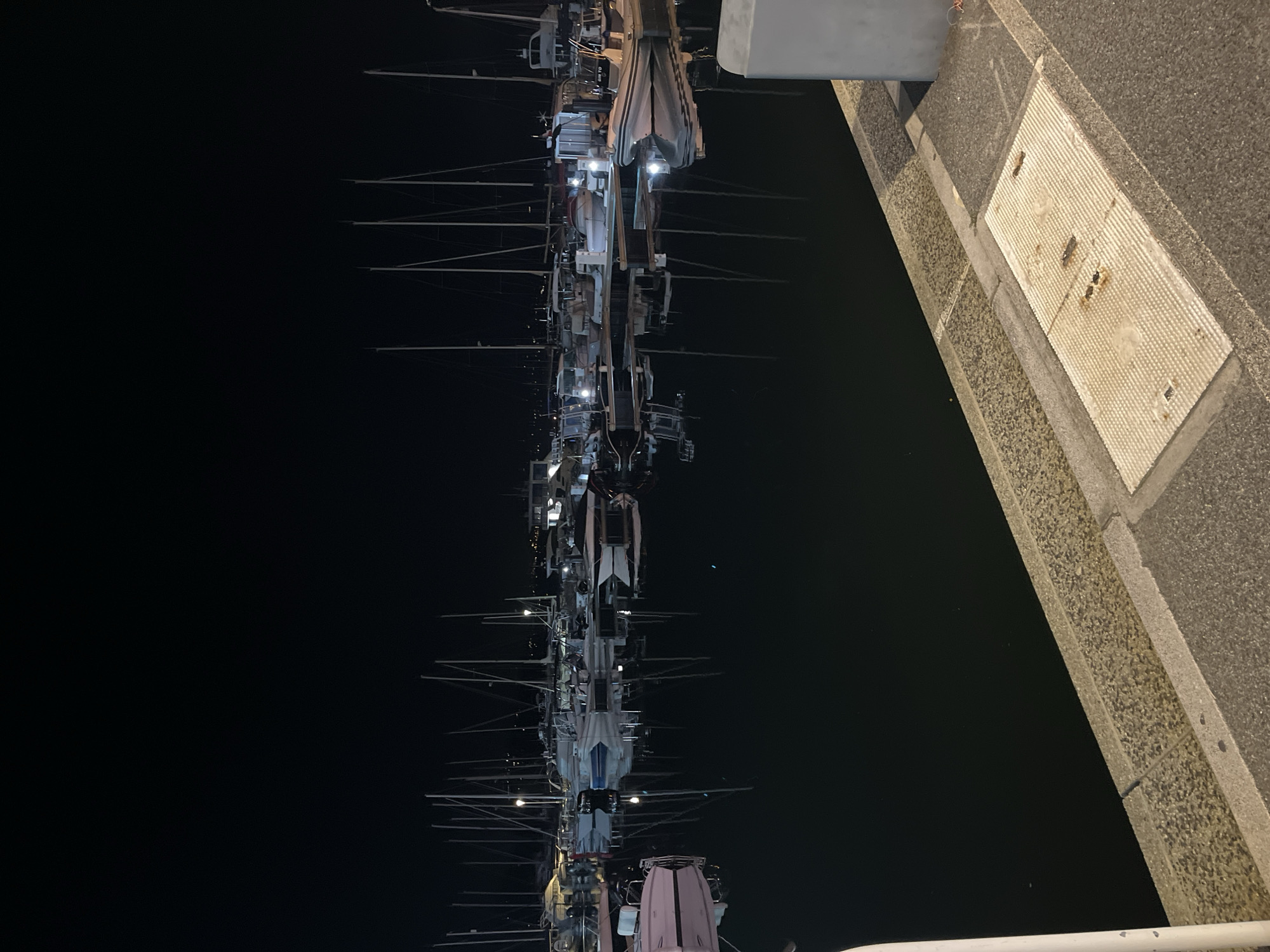 The image shows a night scene at the port of Cavalaire-sur-Mer, France. The view is from a low angle, possibly from a dock or walkway. The photo features yachts moored in the harbor, their masts reaching into the dark night sky. Some yachts have lights on, illuminating parts of their decks. The water reflects these lights, creating a shimmering effect. On the right side of the image, there is a concrete wall or structure, possibly part of the port infrastructure, with a metallic cover or plate. The overall atmosphere is calm and dark, characteristic of a quiet night at the marina.