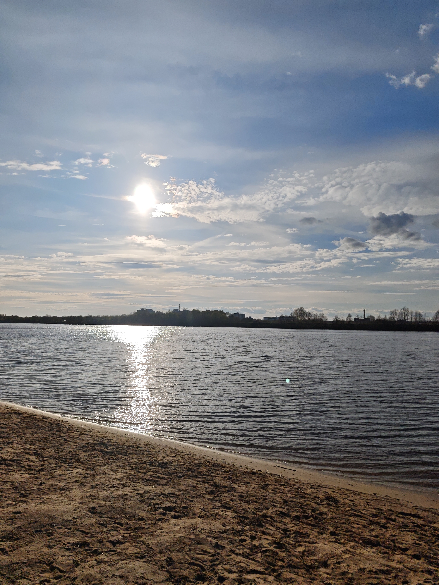 A wide-angle, daytime shot shows a tranquil scene with a body of water, likely a lake or wide river, dominating the frame. The foreground features a sandy beach extending from the bottom left to the mid-right, with distinct textures and variations in color indicative of dry and damp sand near the waterline.
The water surface is rippled, reflecting sunlight in a bright, diffused path leading towards the sun in the sky. The sky is filled with clouds ranging from wispy and thin to more dense formations, suggesting partly cloudy conditions. The sun is visible but partially obscured by clouds, creating a soft, diffused light across the scene.
In the background, across the water, there's a distant shoreline with trees and what appear to be buildings or structures that suggest a city or town skyline. The specific city and any identifiable subjects or people are unidentifiable in the image.