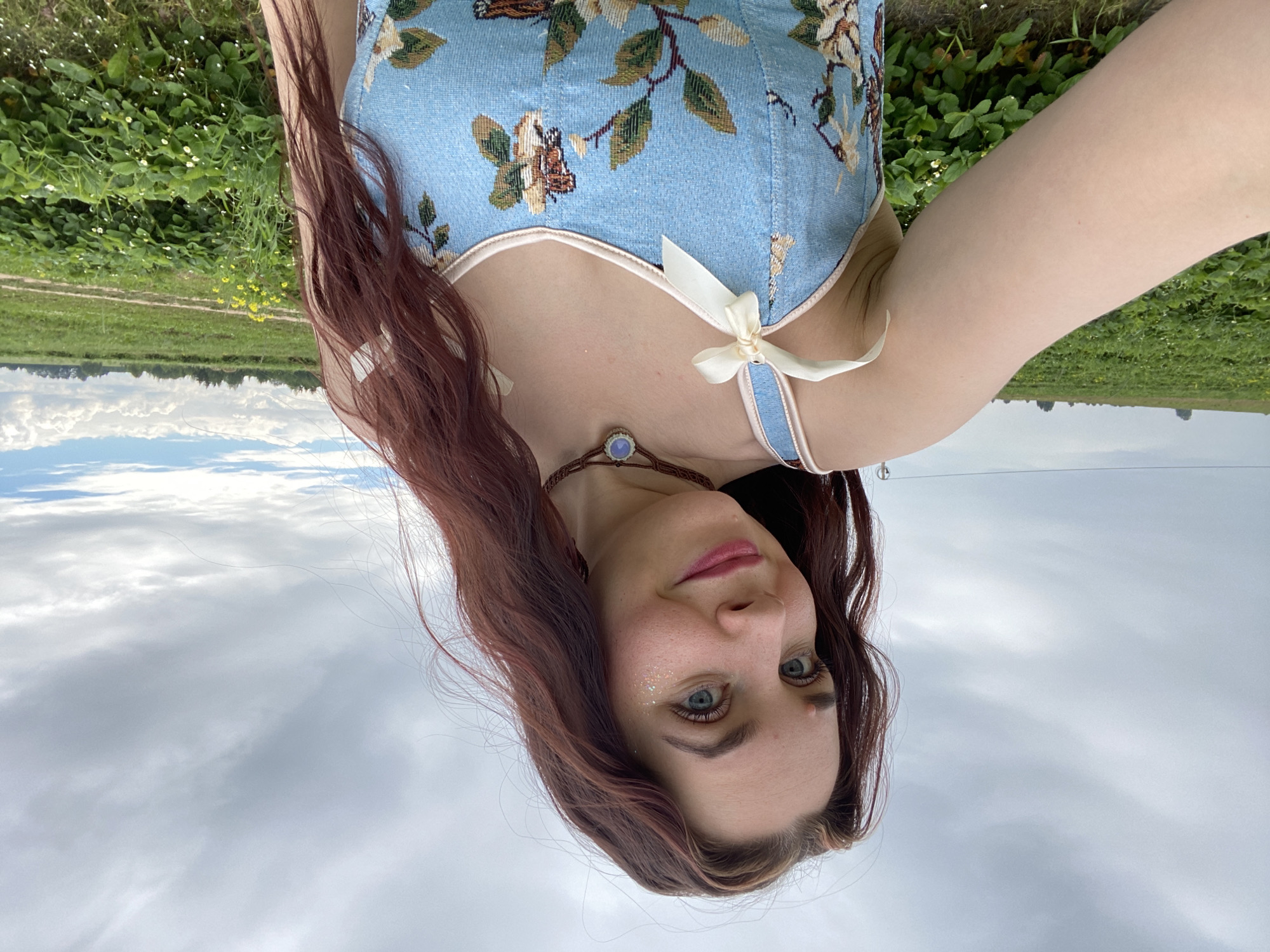 This is a portrait-style, low-angle, medium shot of a young woman posing outdoors. She is oriented with her head towards the bottom of the frame, and her body extending upwards. The woman has long, auburn hair that falls over her shoulders. She is wearing a light blue, patterned corset top with floral and butterfly motifs. A light-colored ribbon is tied in a bow on the left strap of the corset. She has blue eyes, and her face is tilted slightly upwards, looking towards the camera. She is wearing subtle makeup, including glitter on her eyelids and pink lipstick. A necklace with a blue pendant is visible around her neck.
The background is a natural landscape, with a field of green foliage and some yellow flowers to the left of the woman. Further in the background, there is a line of trees and a cloudy sky. The overall impression is that of a warm, overcast day. The sky dominates the upper portion of the frame, with soft, diffused light.
The image appears to have been taken in Foteni, Russia. The combination of the natural setting and the woman's attire suggests a casual, perhaps leisurely activity. There is no visible text in the image.