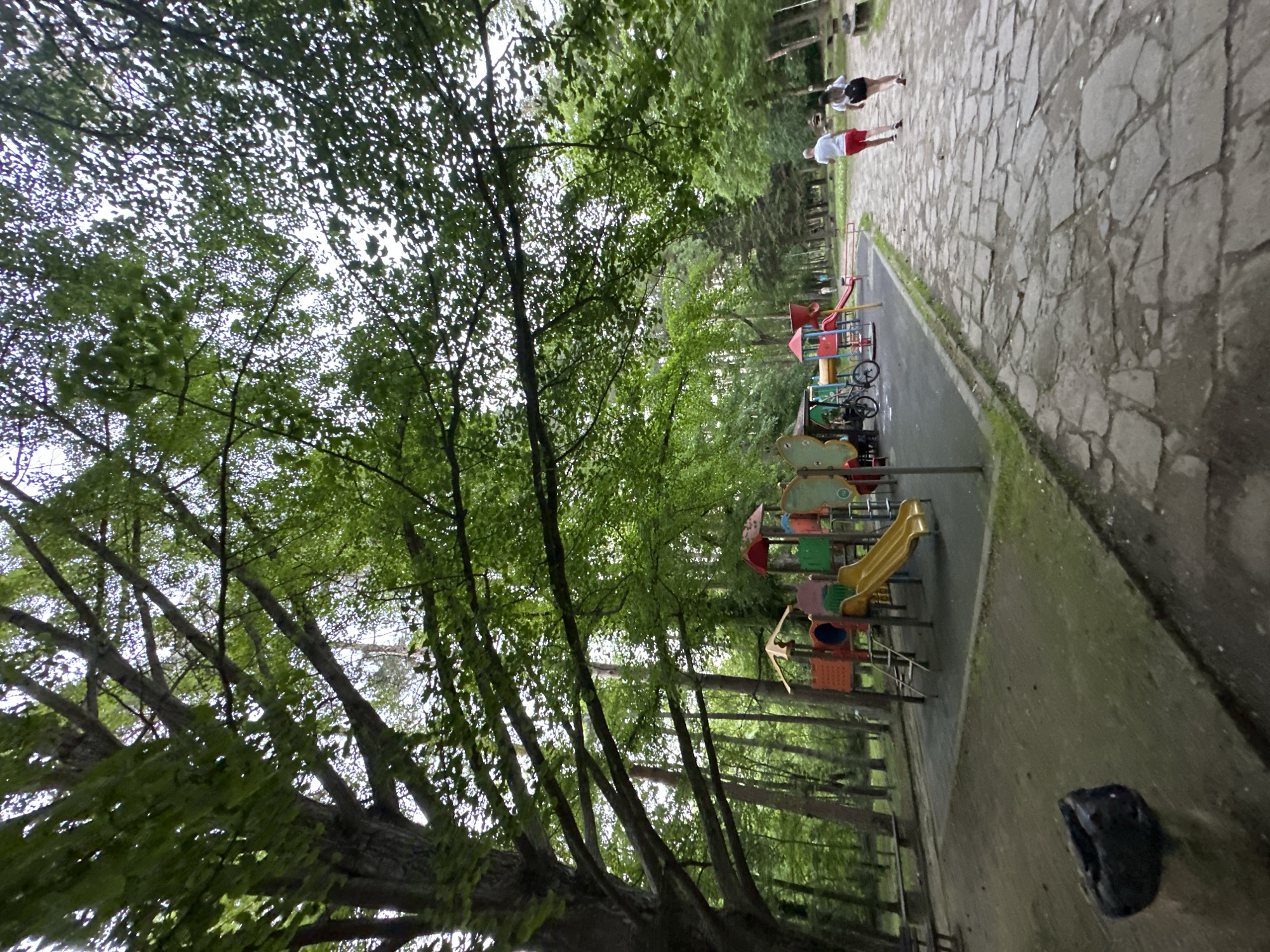 The image depicts a park scene in Lovech, Bulgaria. A stone pathway extends from the upper right towards the center. Two people, one wearing red shorts and a white shirt and the other wearing black shorts and a white shirt, are walking along the path. A playground with slides, swings, and other colorful equipment is visible adjacent to the path. The left side of the image is dominated by trees with green leaves. A dark-colored object rests on the ground in the lower right corner.