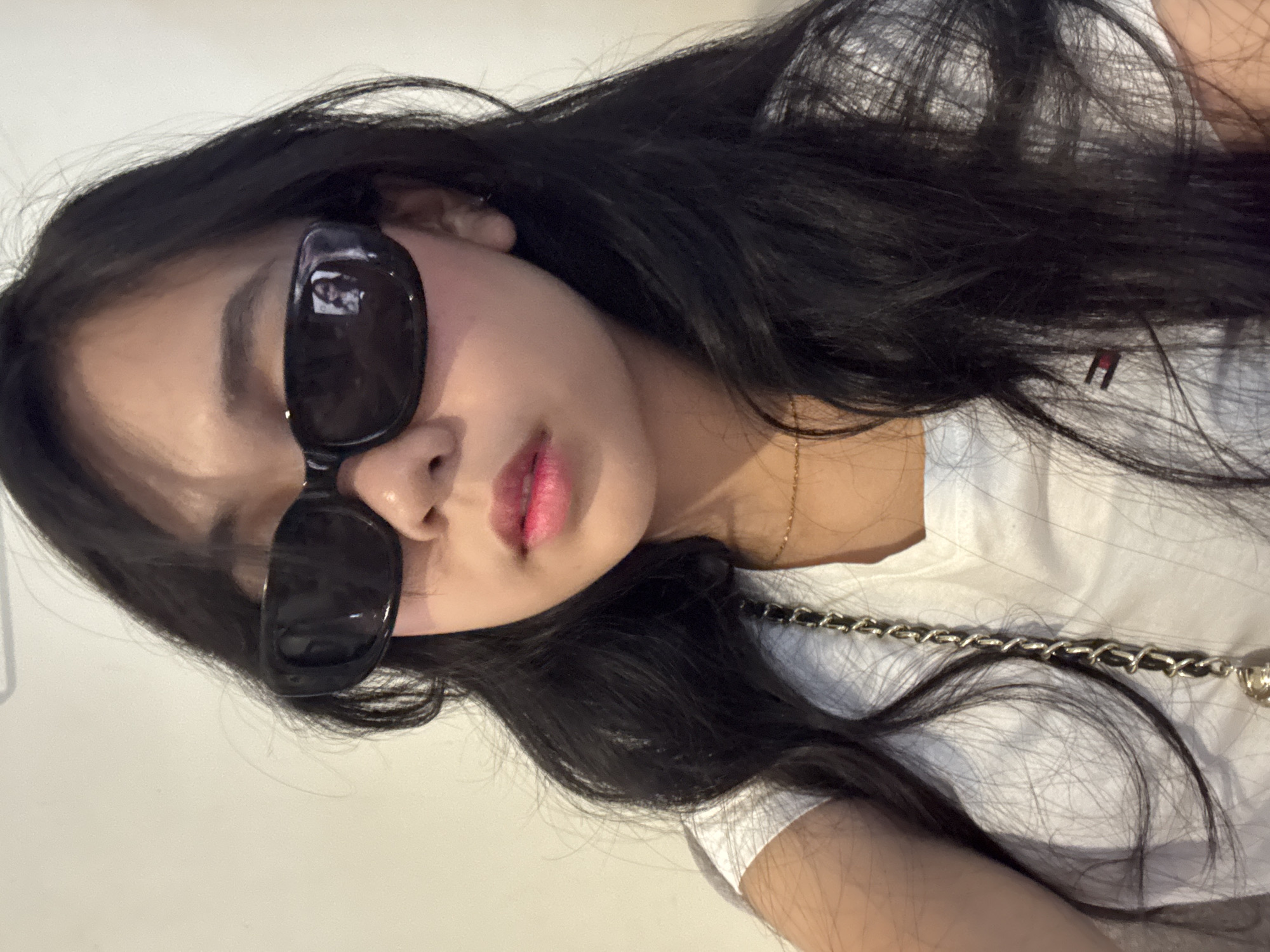 A young woman with long, dark hair and dark sunglasses is captured in a close-up, frontal shot. She is wearing a white t-shirt adorned with a small, red, white, and blue rectangular logo on the left side. A gold chain necklace is visible around her neck, and a black and silver chain strap from a purse rests on her shoulder. Her lips are painted a bright pink. The background is a plain, light-colored wall. The reflection in her sunglasses shows another person, possibly the photographer. The scene appears to be indoors, in an urban setting like Davao City, Philippines, possibly in a residential or commercial space given the simple background.