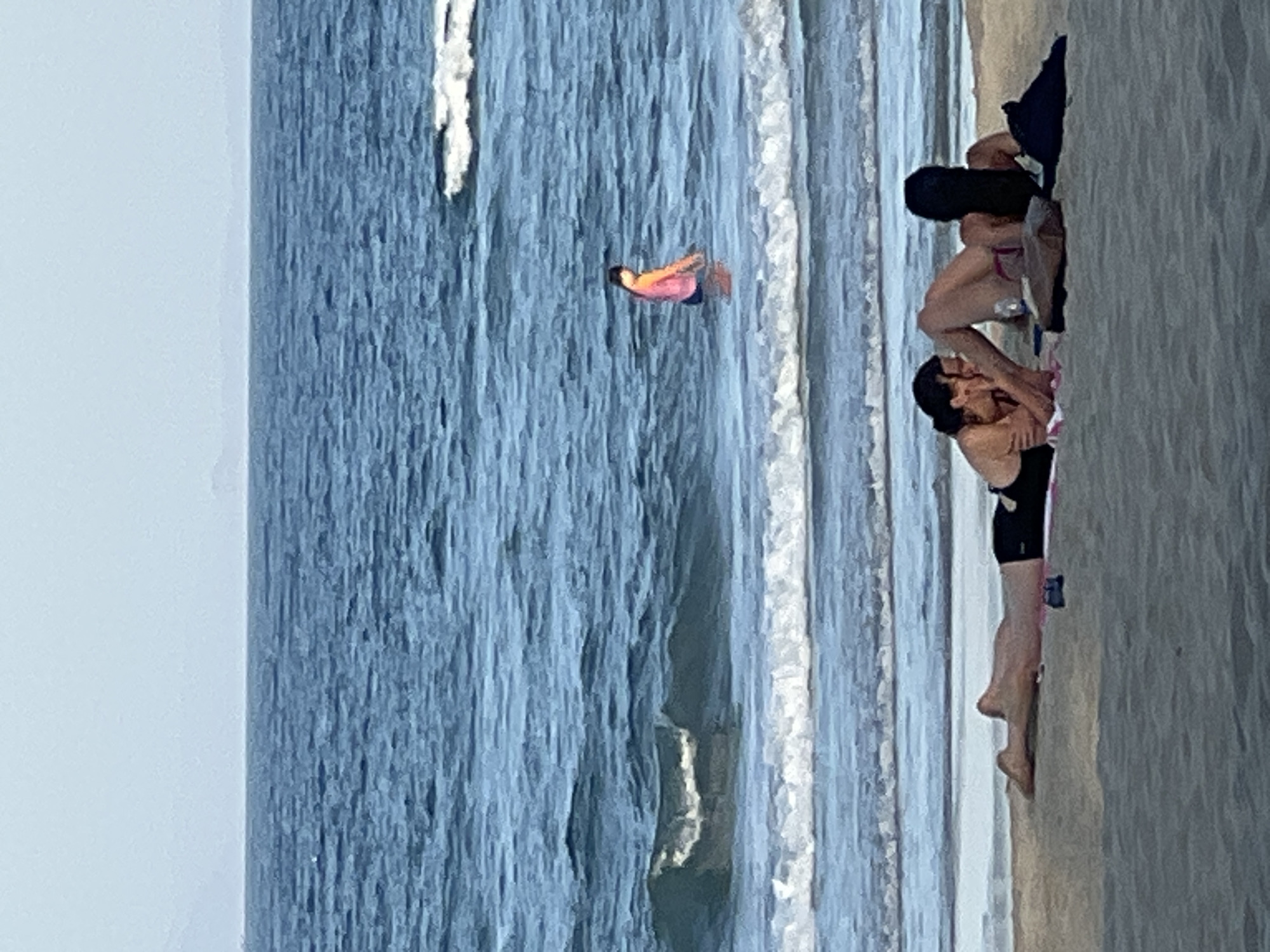Two individuals are lying on a sandy beach in Đà Nẵng, Vietnam, near the ocean. One person is on their stomach, facing the water, wearing a black swimsuit. The other person is lying on their back, facing away from the water, with their legs extended. They are both on towels. In the background, a person in a pink shirt and blue bottoms is standing in the water. The ocean is blue with white waves. The sky is a light blue.
