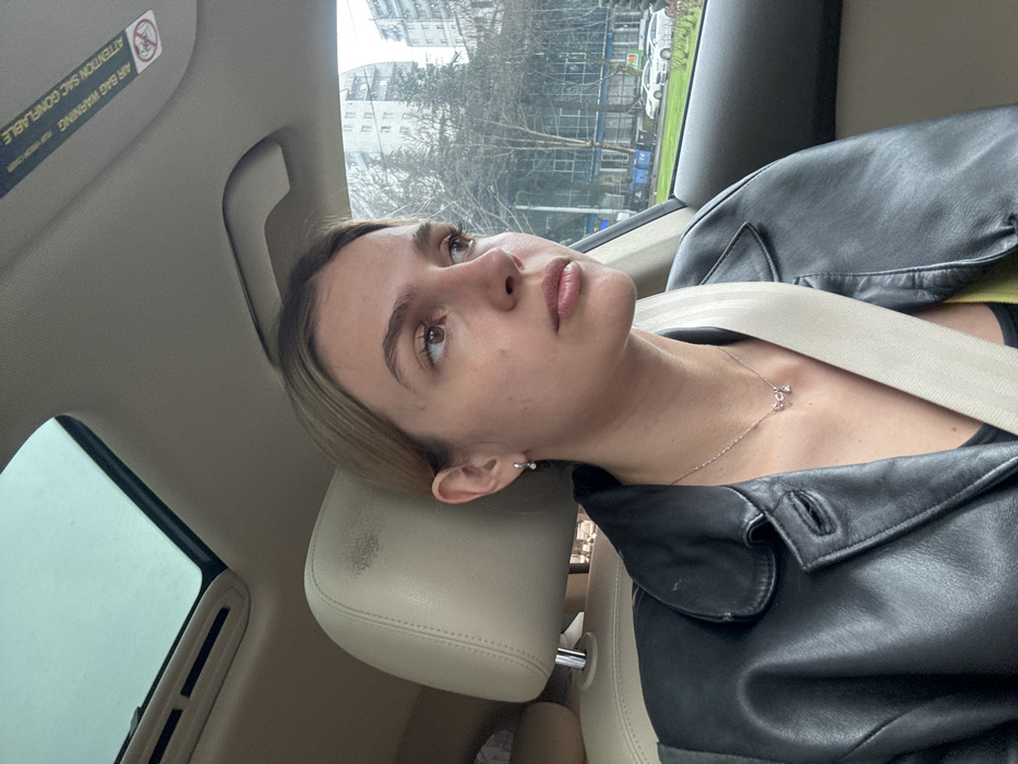 A female individual is seated in the passenger seat of a vehicle, positioned at a slight angle, looking towards the right out the side window. She has light-colored hair, pulled back, and wears a black leather jacket over an undershirt, with a light-colored seatbelt diagonally across her torso. A thin silver necklace with a small, reflective pendant adorns her neck, and a small silver-colored hoop earring with a clear stone is visible on her left ear. The vehicle's interior features a beige headrest and ceiling, which includes a sunroof revealing an overcast sky and a warning sticker for "AIR BAG WARNING ATTENTION SAC GONFLABLE."
The external environment visible through the window identifies an urban streetscape in Tbilisi, Georgia. The scene features deciduous trees with minimal foliage, suggesting a colder season. In the mid-ground, a commercial building with large glass panels and blue waste receptacles is visible. Beyond this, multiple multi-story residential buildings are present. Several white passenger vehicles are parked along the street.