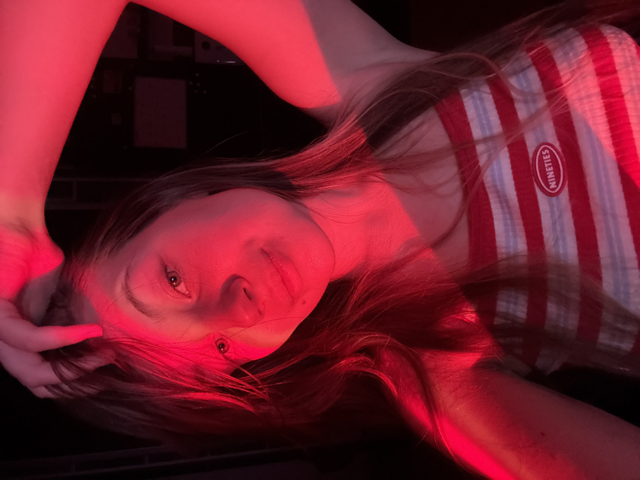 A young adult female is depicted in a medium close-up, illuminated by strong red light within an otherwise dark, indistinct indoor environment, located in the city of Vozdovac Urban Municipality, Serbia. She has long, light brown hair, which she is touching with her right hand, fingers splayed, above her forehead. She is looking directly at the viewer with a slight, closed-mouth smile. She wears a sleeveless top with horizontal red and white stripes. An oval-shaped red patch with the word "NINETIES" in white text is visible on the upper chest area of her top. Her left arm is partially visible on the right side of the frame. The red light casts shadows, particularly across the left side of her face and torso.
