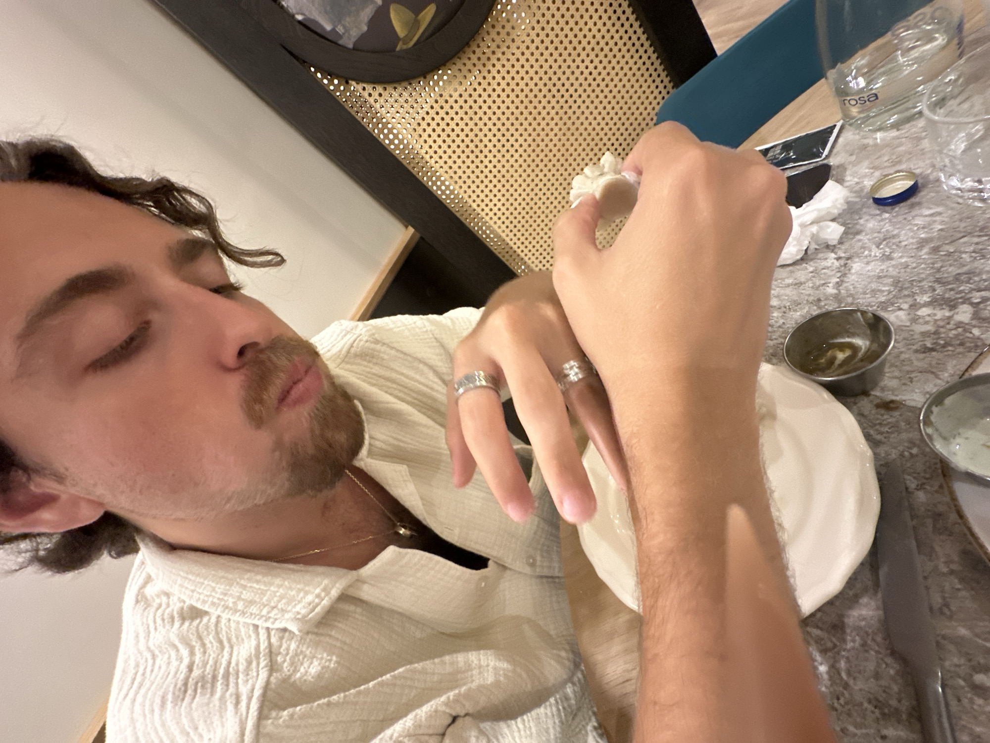 In this close-up, indoor shot, a person with dark, curly hair and a light-colored, textured shirt is seen from the chest up, leaning forward and looking down at their hands, which are actively working with a ball of white dough on a speckled, gray countertop. The person wears a thin gold chain with a pendant and two rings on their right hand. To the right of the dough, a small metal bowl with green liquid is visible, along with a crumpled white paper napkin, a blue and silver cap, and two clear glasses, one of which has "rosa" printed on it. In the background, a dark frame with a perforated, tan material and a glimpse of a picture or object is present. The lighting appears warm and ambient. The scene is consistent with a restaurant or domestic setting where food preparation is taking place.