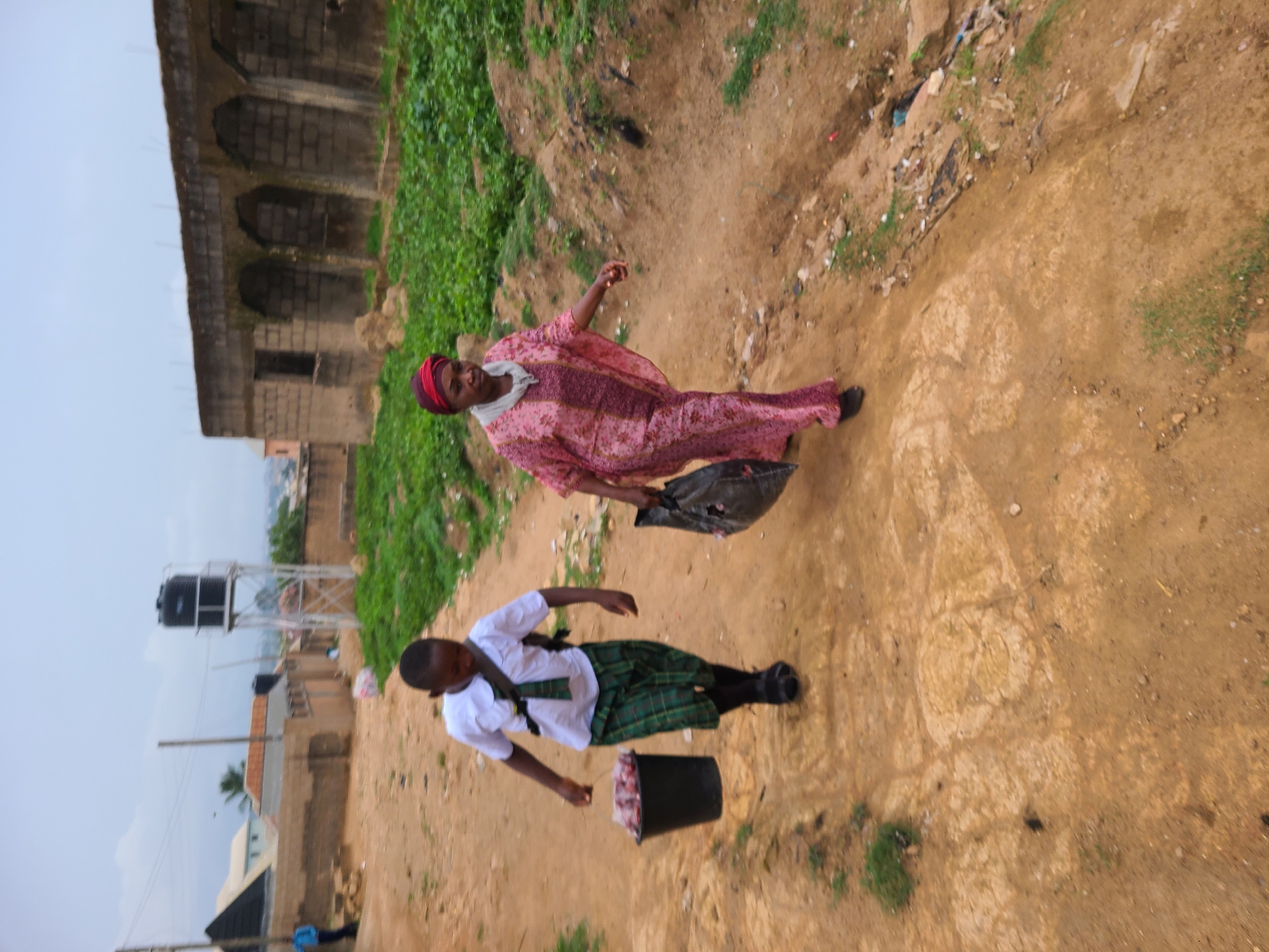 Two individuals are walking on a dirt path. The first is a woman, wearing a red and pink patterned dress with a matching head covering. She is carrying a black plastic bag. The second individual appears to be a student in a green and white uniform with a tie. The student is also carrying a black bucket filled with an unidentifiable pink material.
The background includes a partially visible building with arched windows and a water tower structure. A patch of green vegetation can be seen behind the woman, and general ground debris is scattered along the path. The scene is located in an urban or semi-urban area in Jos, Nigeria.