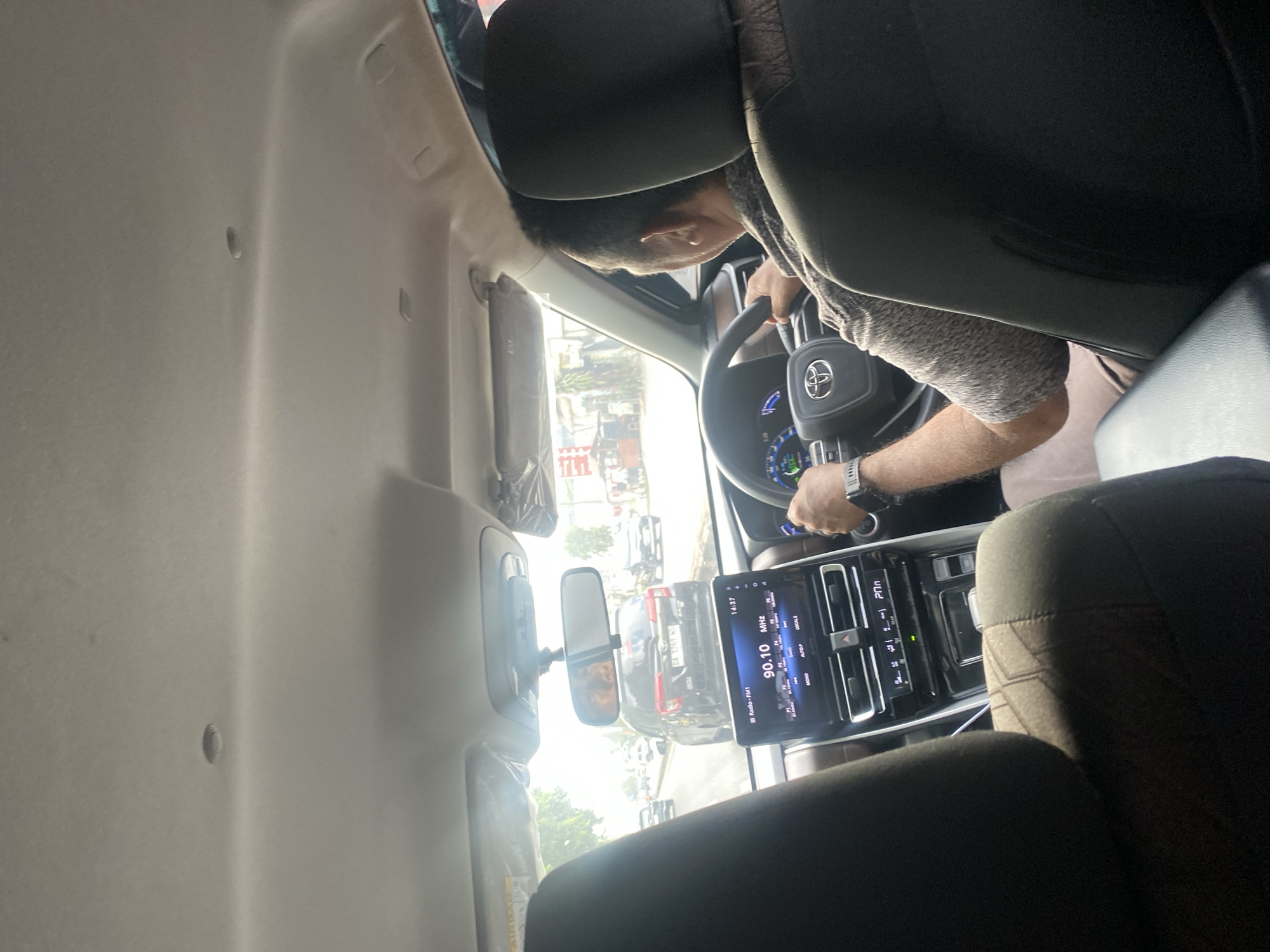 The image shows the interior of a car, viewed from the back seat, looking forward. A man is driving the car. He is wearing a grey patterned short-sleeved shirt and appears to be of Indonesian ethnicity, possibly in his 40s or 50s. He is holding the steering wheel, which has the Toyota logo on it. The car's dashboard is visible, with a touchscreen display showing radio information and climate control settings. The car is on a road in Banjarbaru, Indonesia, with other vehicles visible outside. The time on the dashboard indicates 14:37. The overall scene depicts a common daily activity of driving in a city environment.