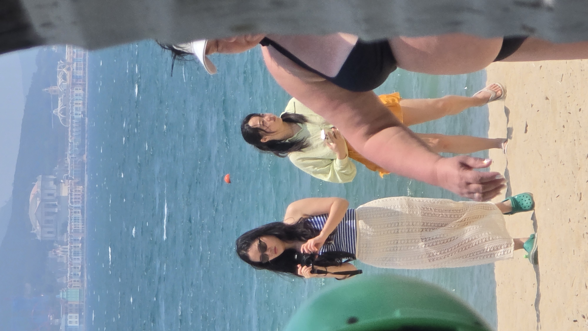 The image captures a scene at a beach in Nha Trang, Vietnam. In the background, a coastline with buildings is visible. The turquoise water of the ocean is calm. In the foreground, there are three individuals. The closest person to the camera is partially obscured, but is wearing a black bikini bottom and a white visor. There are two women standing on the sand. One woman is wearing a blue and white striped top with a long white lacy skirt and green Crocs. She has sunglasses on and is holding a camera, possibly taking a photo. The other woman is wearing a light green, long-sleeved shirt and short orange skirt. She is holding something in her hand and is smiling. A small orange ball or object floats in the water between the two women.