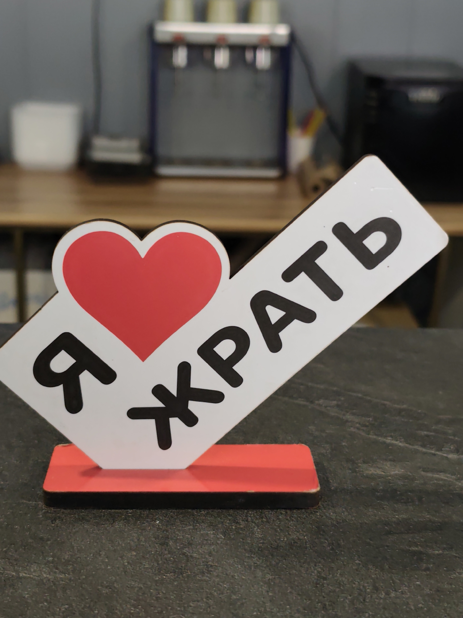 A close-up photograph shows a table in what appears to be a public space. On the table is a sign that reads "Я ХРАТЬ" with a heart symbol. The words are written in black on a white background with a red heart. The sign is placed on a red and black base. Behind the sign are blurred objects including a coffee machine, a white container, and a wooden counter. The location is assumed to be Chaikovsky, Russia based on the Cyrillic text on the sign.