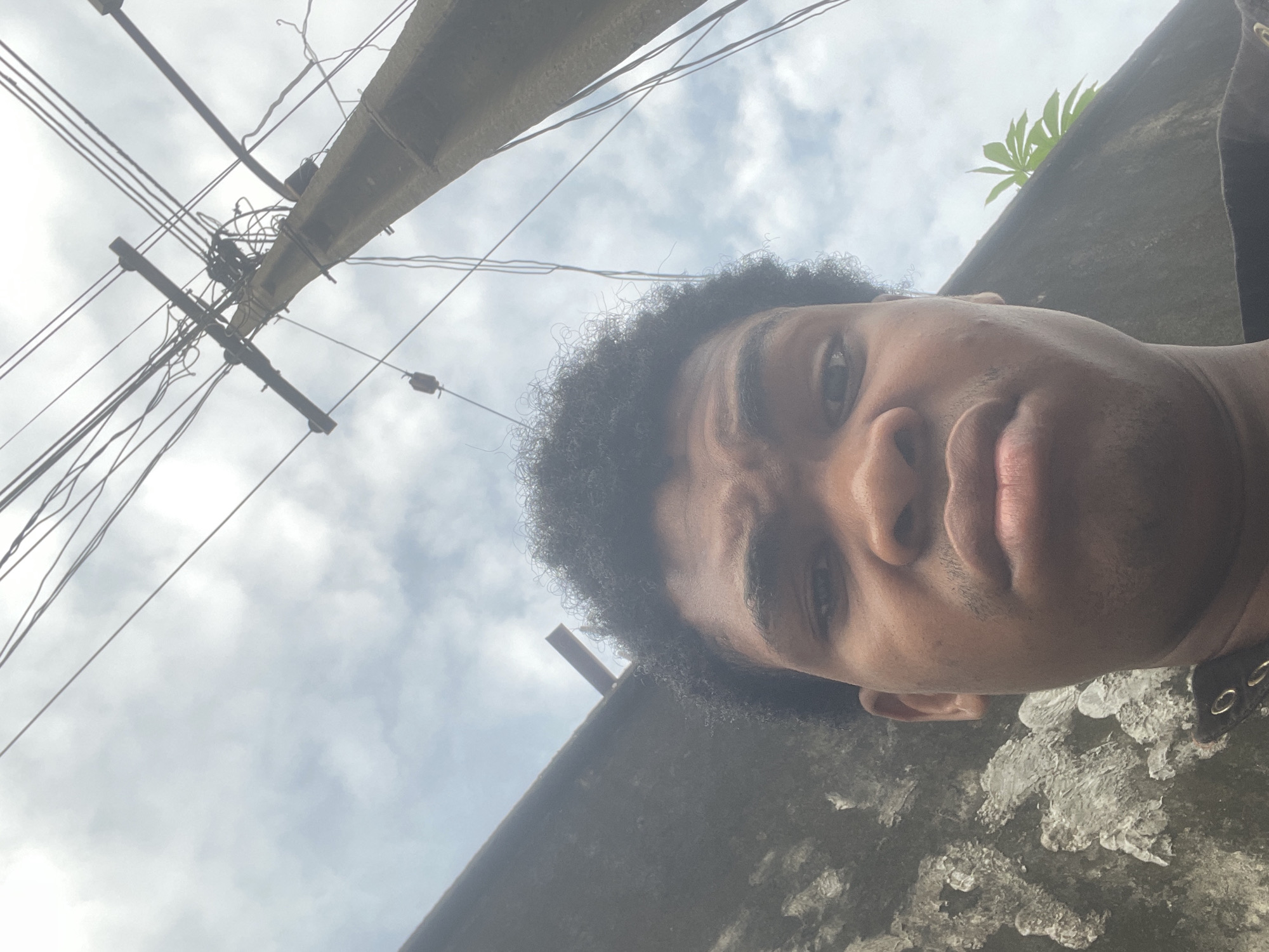 A close-up, low-angle selfie taken outdoors, likely in Ado, Nigeria. The subject is a young Black male with curly dark hair, looking directly at the camera with a neutral expression. His face is the primary focus, occupying the lower half of the frame. Behind him, a concrete utility pole with numerous tangled wires extends upwards, crisscrossing against a cloudy sky. To the left, a textured, dark grey wall runs along the bottom of the frame. A plant with green leaves is visible to the right, partially obscuring the sky behind the utility pole. The lighting suggests an overcast day.