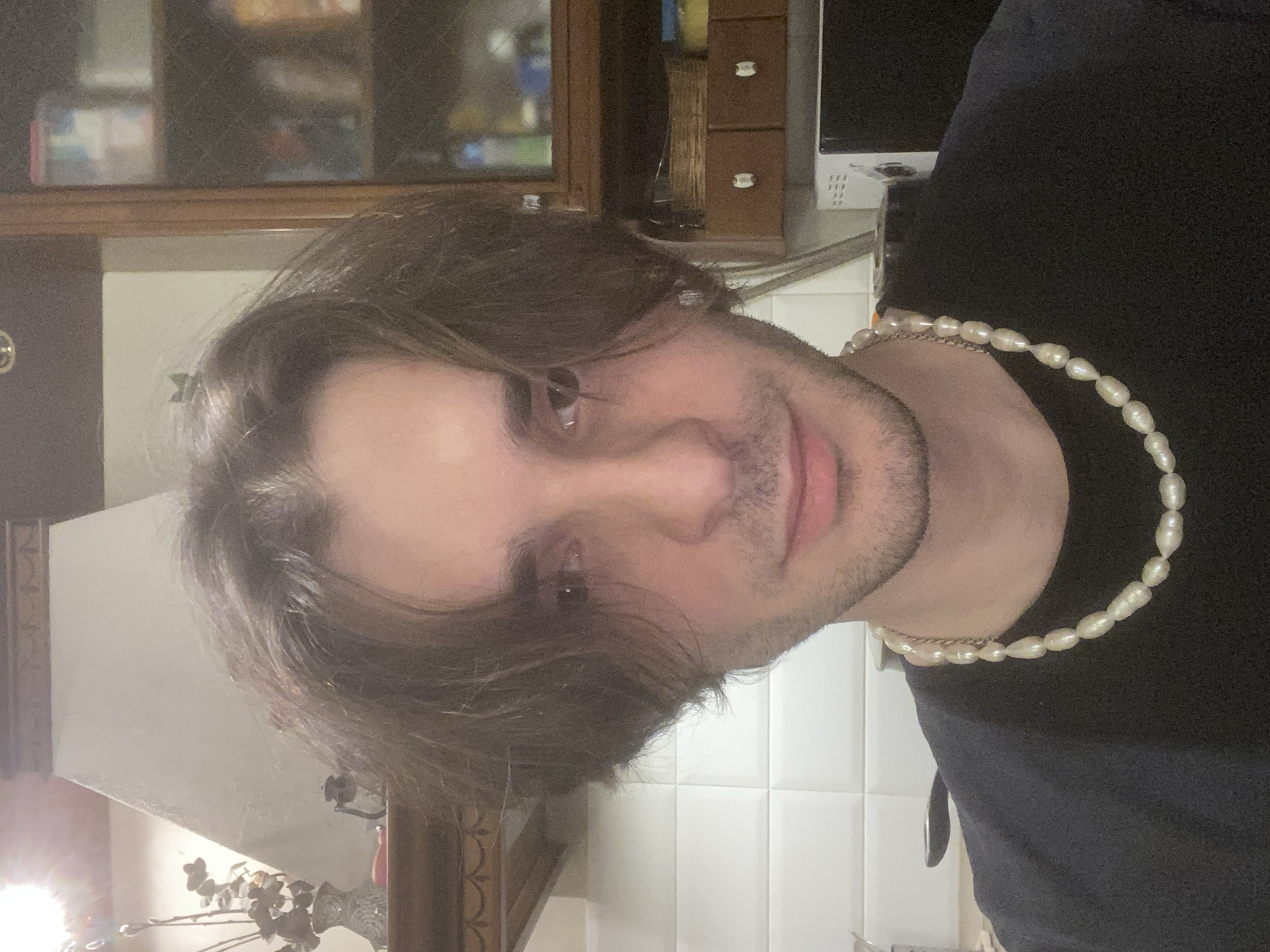 A close-up, slightly angled, indoor photograph captures a fair-skinned person wearing a dark t-shirt and a pearl necklace. The person has dark, medium-length hair and stubble. The background includes a brown wooden cabinet, a microwave, and white tiled walls. The location within Moscow, Russia cannot be determined from the available visual information.
