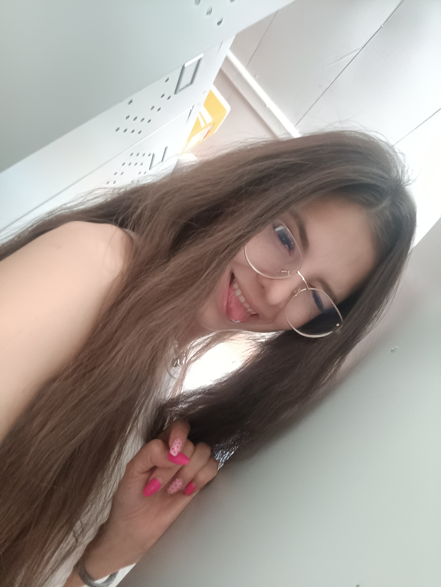 A young woman with long brown hair and glasses smiles and sticks out her tongue while posing for a photo. She is wearing pink nail polish with a polka dot design on some nails. Her left hand is visible, adorned with a silver ring and a bracelet. She appears to be leaning against a white wall, with a perforated metal panel above her. The location is Прикамский, Russia.