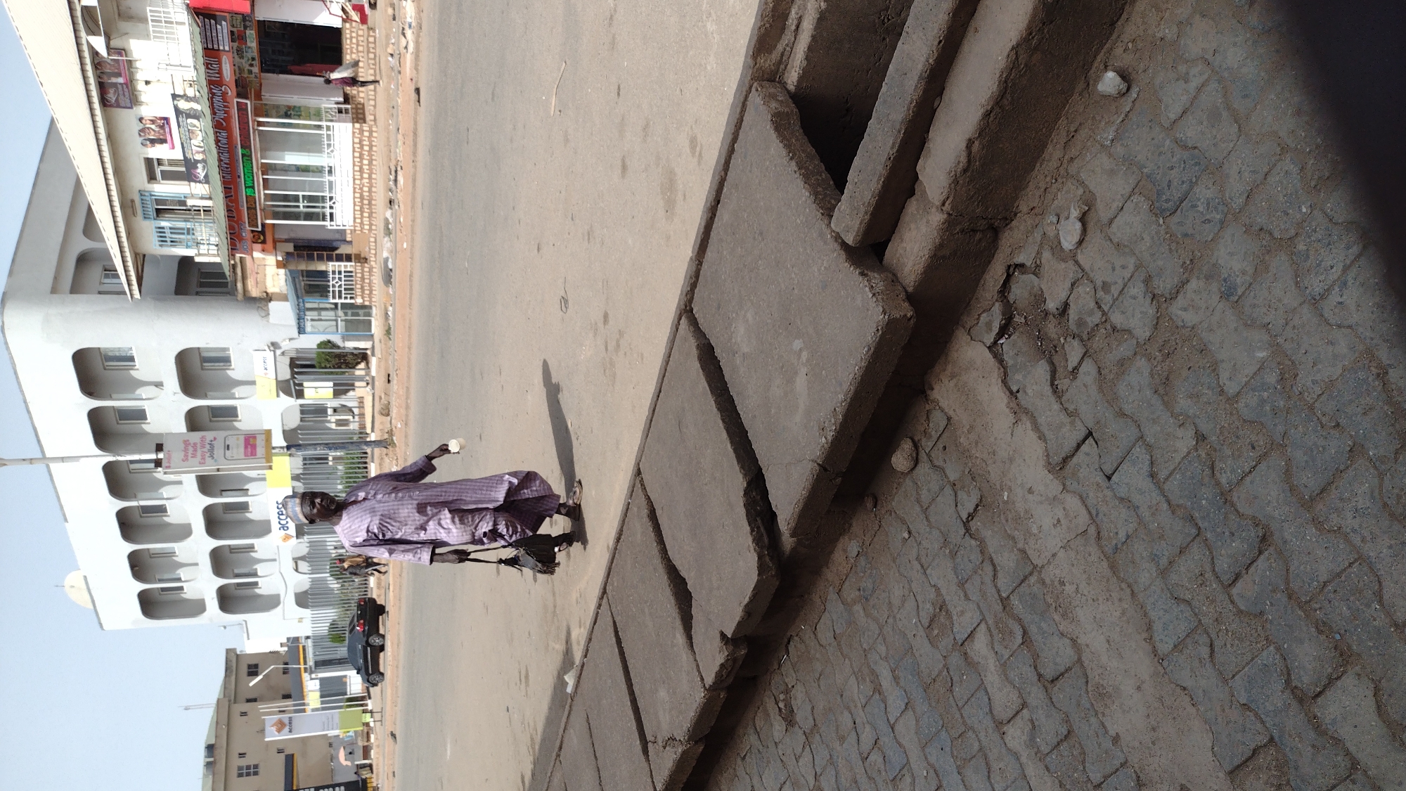 An elderly man in a purple robe walks along a street in Jos, Nigeria. He is carrying a cup in one hand and a walking stick in the other. The street is paved and there is a curb with concrete covers over a drainage system. On the other side of the street are buildings with businesses and apartments. There are also street signs visible. The day is sunny.