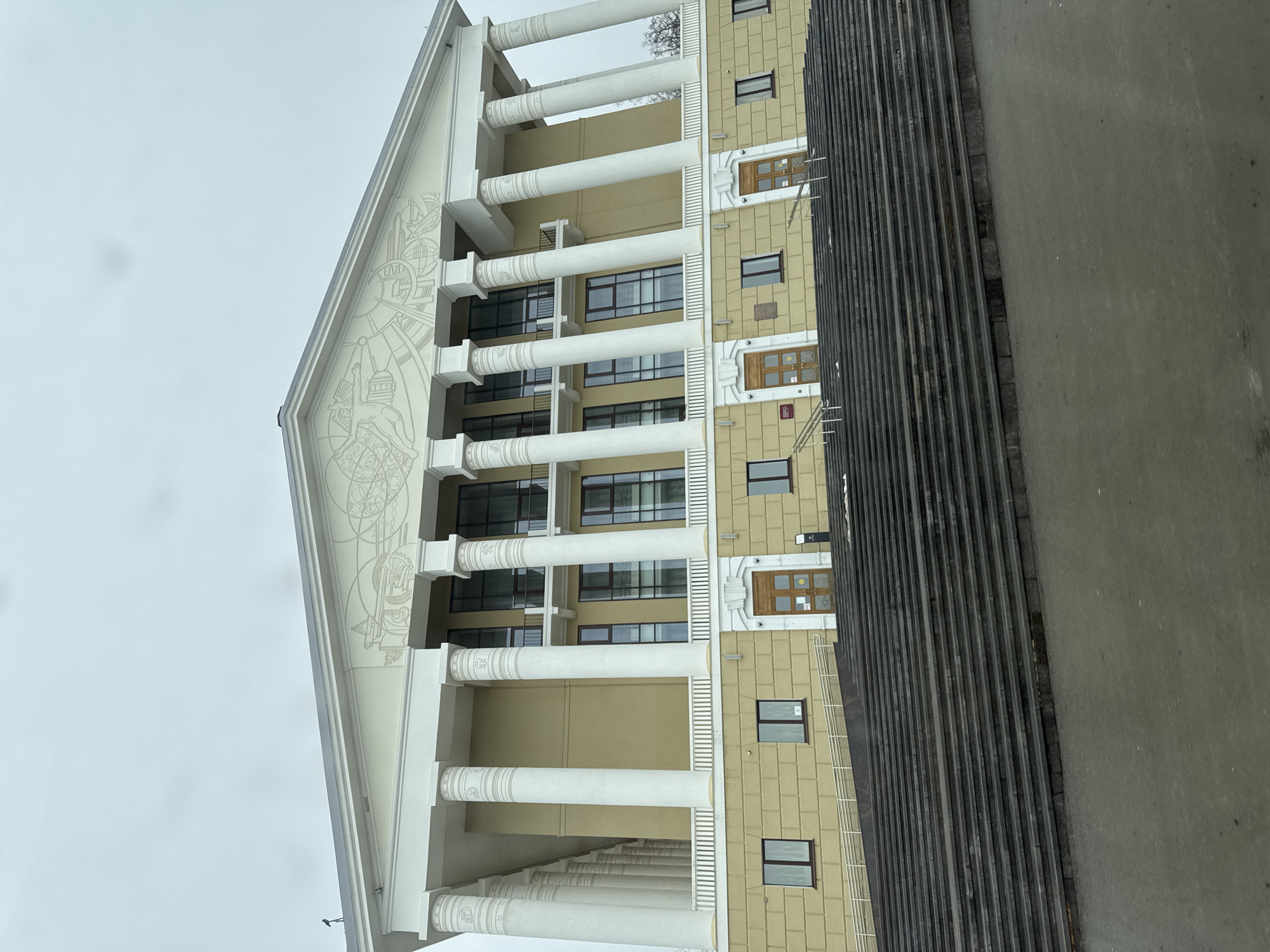 A low-angle, slightly rotated photograph captures a building with distinct architectural features. The facade includes several rows of windows, some flanked by decorative doors, set in a grid-like pattern on a yellow-tan wall. Flanking this section are large, white columns with patterned supports, reaching from the ground to the top of the building. Above the columns is a triangular pediment adorned with a detailed, light-colored graphic design. To the right, partially obscuring the building, are linear, vertical elements that appear to be part of an adjacent structure. The sky is overcast. This is the building of the administration of the "Votkinsky Zavod" located in Votkinsk, Russia.