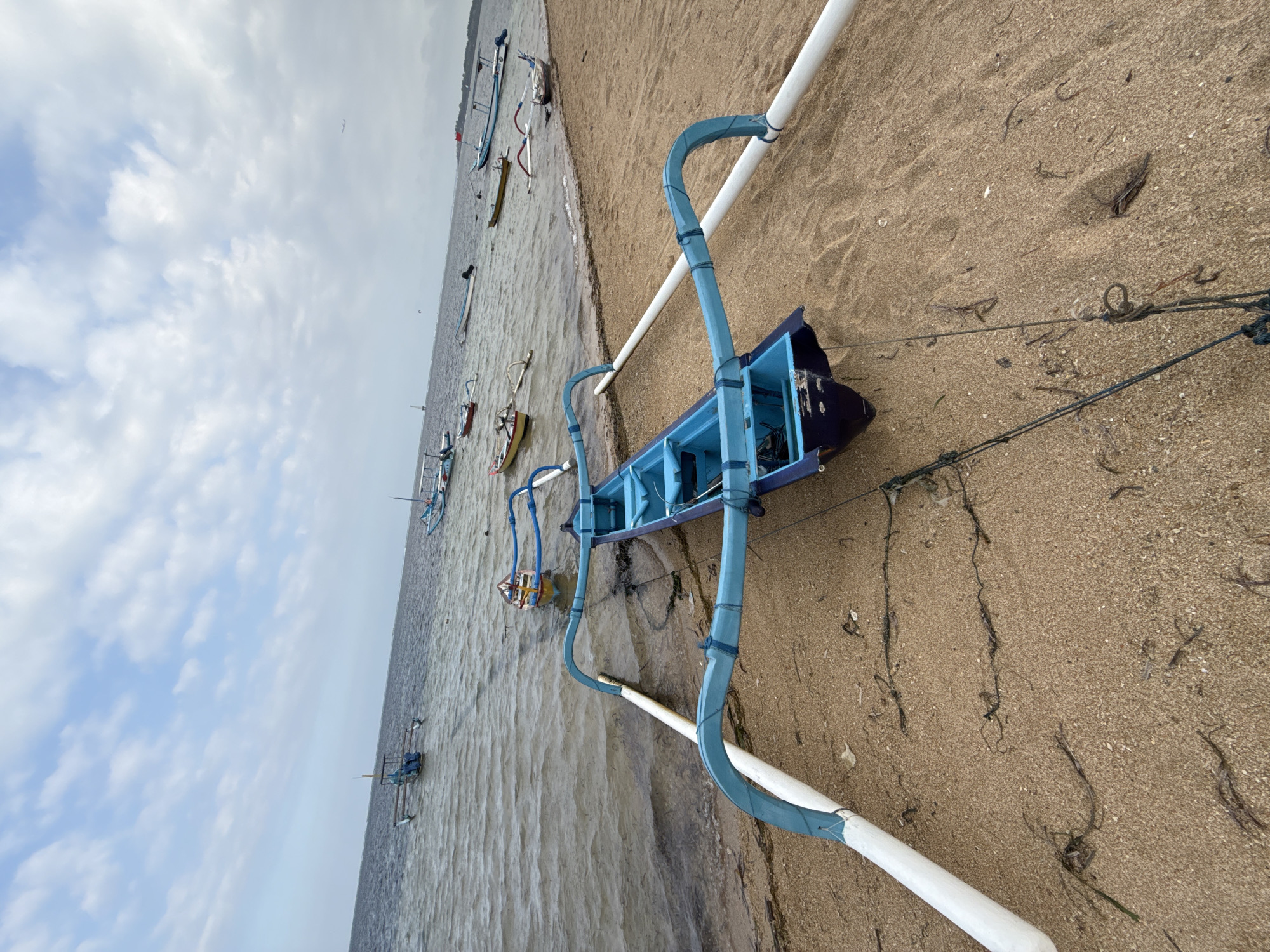 The image shows a beach scene with several outrigger canoes, typical of those used in Denpasar, Indonesia. In the foreground, a blue outrigger canoe rests on the sandy shore, its outriggers extending outwards. The water is calm, with several other canoes scattered across the surface. The sky is partly cloudy, with a few wispy clouds against a pale blue backdrop. The sand is light brown and appears to be mixed with small pieces of seaweed and debris. The overall impression is a serene coastal landscape, likely a popular spot for local fishermen or tourism in Denpasar.
