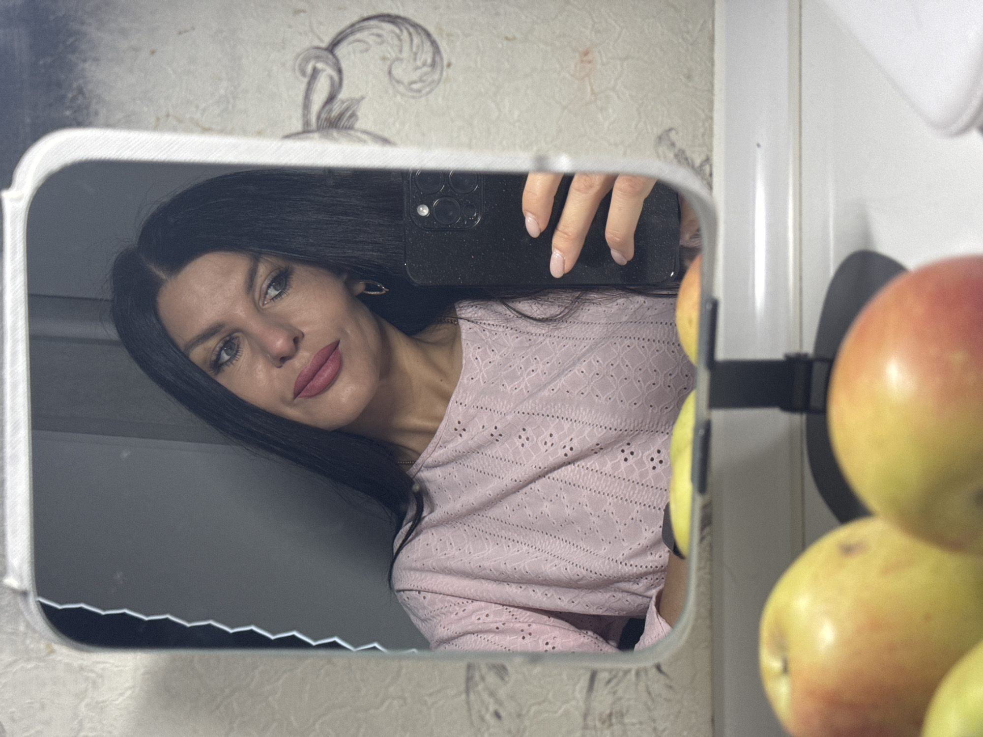 A woman is taking a selfie in a mirror. The woman has long black hair, fair skin, light-colored eyes, and is wearing a pink long-sleeved shirt. She is holding a black smartphone in her right hand. To the right of the mirror, several apples are visible in a fruit holder. The wallpaper behind the mirror is patterned. The photo appears to be taken in an indoor environment, possibly a kitchen, in a location in the city of Chaikovsky, Russia.