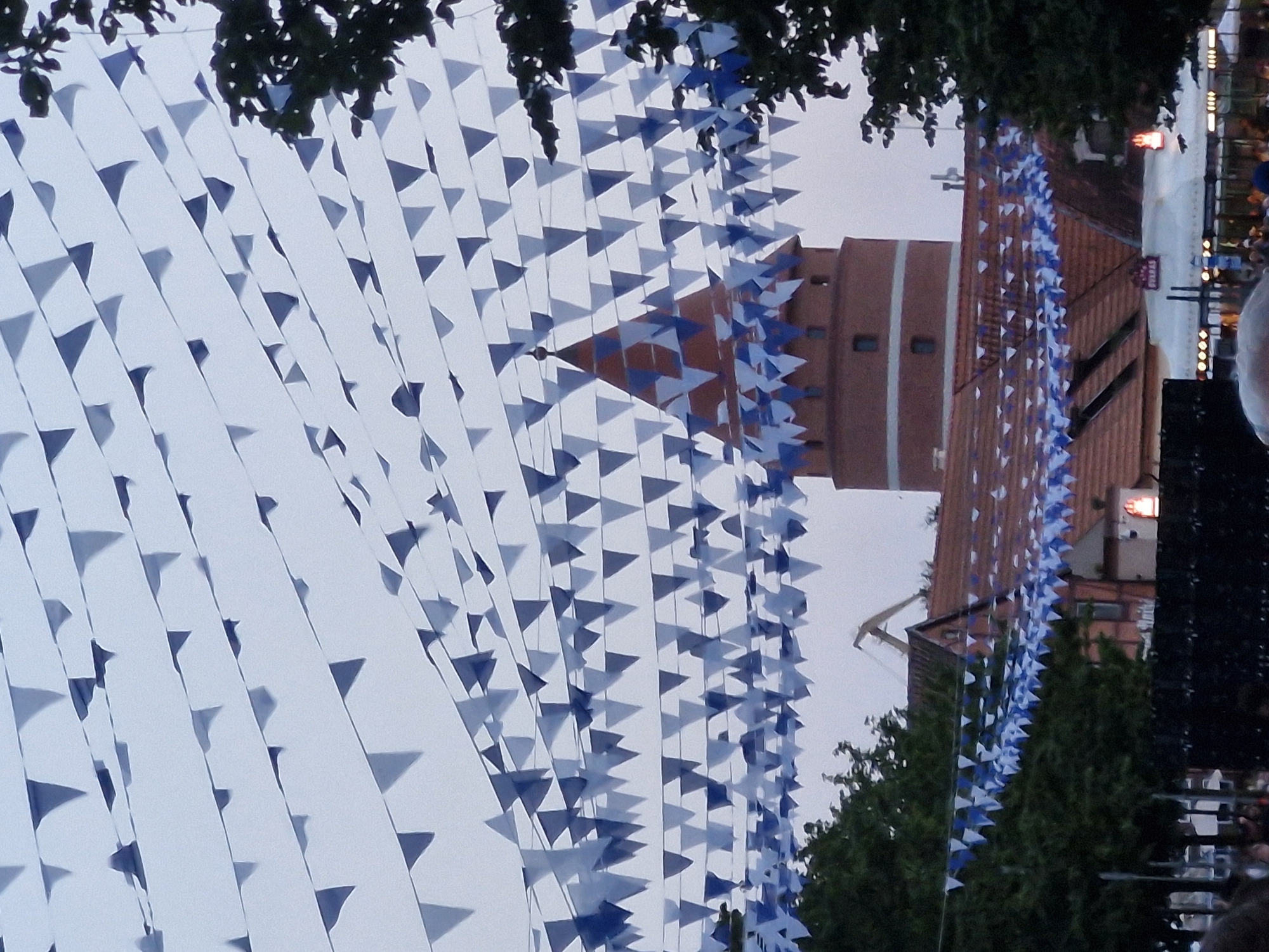 The image shows a festival scene in Klaipėda, Lithuania, with numerous strings of blue and white triangular flags strung across the sky. In the background, a round, red-brick tower with a conical roof, also decorated with flags, is visible. Adjacent to the tower is a building with a red-tiled roof. Trees frame the scene on the left and right. The sky is overcast. Below the buildings, a dark, blurred area suggests a crowd of people.