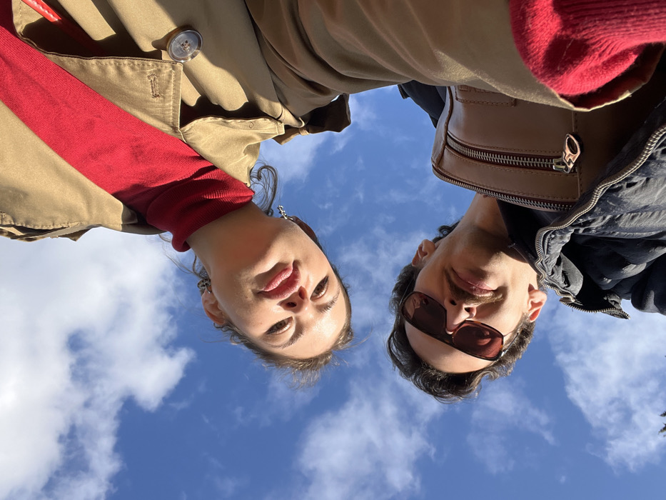 Two individuals are captured in a low-angle photograph against a clear blue sky with scattered white clouds, taken outdoors in the city of Budva, Montenegro. The male subject, positioned higher in the frame, wears a dark puffer jacket, brown-tinted sunglasses, and exhibits a mustache. A tan-colored zippered bag or strap is visible on his left shoulder. The female subject, positioned lower and slightly to the left, wears a red turtleneck sweater and a tan trench coat, with small earrings visible. Her lips are slightly pursed, and both individuals are looking directly downwards towards the camera.