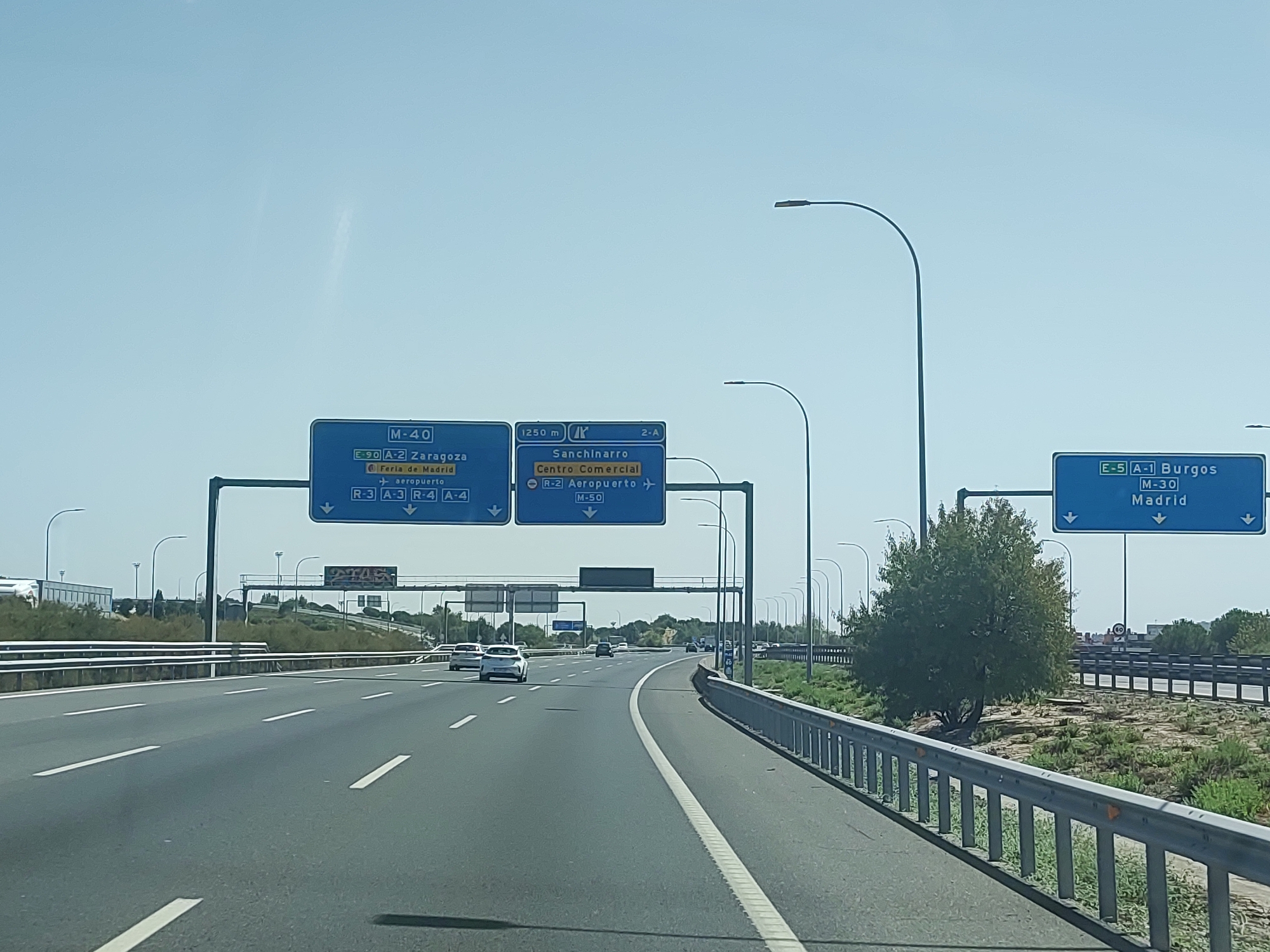 The image shows a highway scene in Madrid, Spain, likely on the outskirts of the city. Several blue highway signs are visible, indicating directions to various destinations. One sign shows "M-40" and directions to "E-90 A-2 Zaragoza", "Feria de Madrid", and "Aeropuerto". Another sign indicates "Sanchinarro", "Centro Comercial", and "R-2 Aeropuerto" with a distance of "1250 m". A third sign on the right side of the highway displays "E-5 A-1 Burgos" and "M-30 Madrid". The highway itself is multi-lane and appears to be an elevated road or flyover. Cars are driving on the highway. The environment includes a clear blue sky, some vegetation along the sides of the road, and streetlights. The scene is captured during the daytime, under bright sunlight.