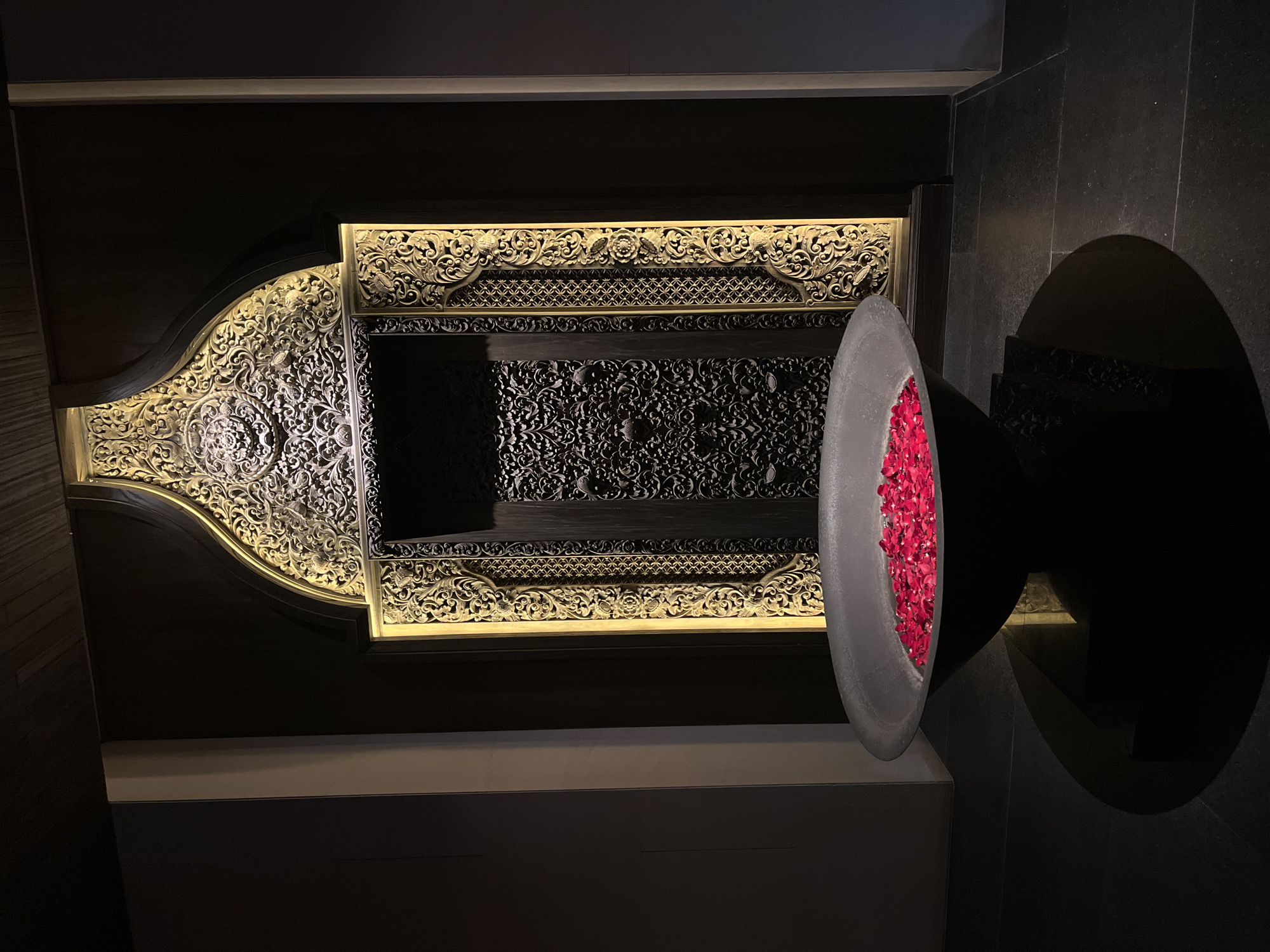 The image depicts an interior scene with a large, dark basin filled with red rose petals in the foreground. The basin rests on a dark, stepped pedestal. Behind the basin is an ornate, arched doorway with intricate carvings and gilded accents. The doorway appears to be made of dark wood, with decorative panels featuring detailed patterns. The overall ambiance is luxurious and dimly lit, suggesting a spa or high-end establishment. The floor is made of dark, rectangular tiles. The location is Kuta Selatan, Indonesia.