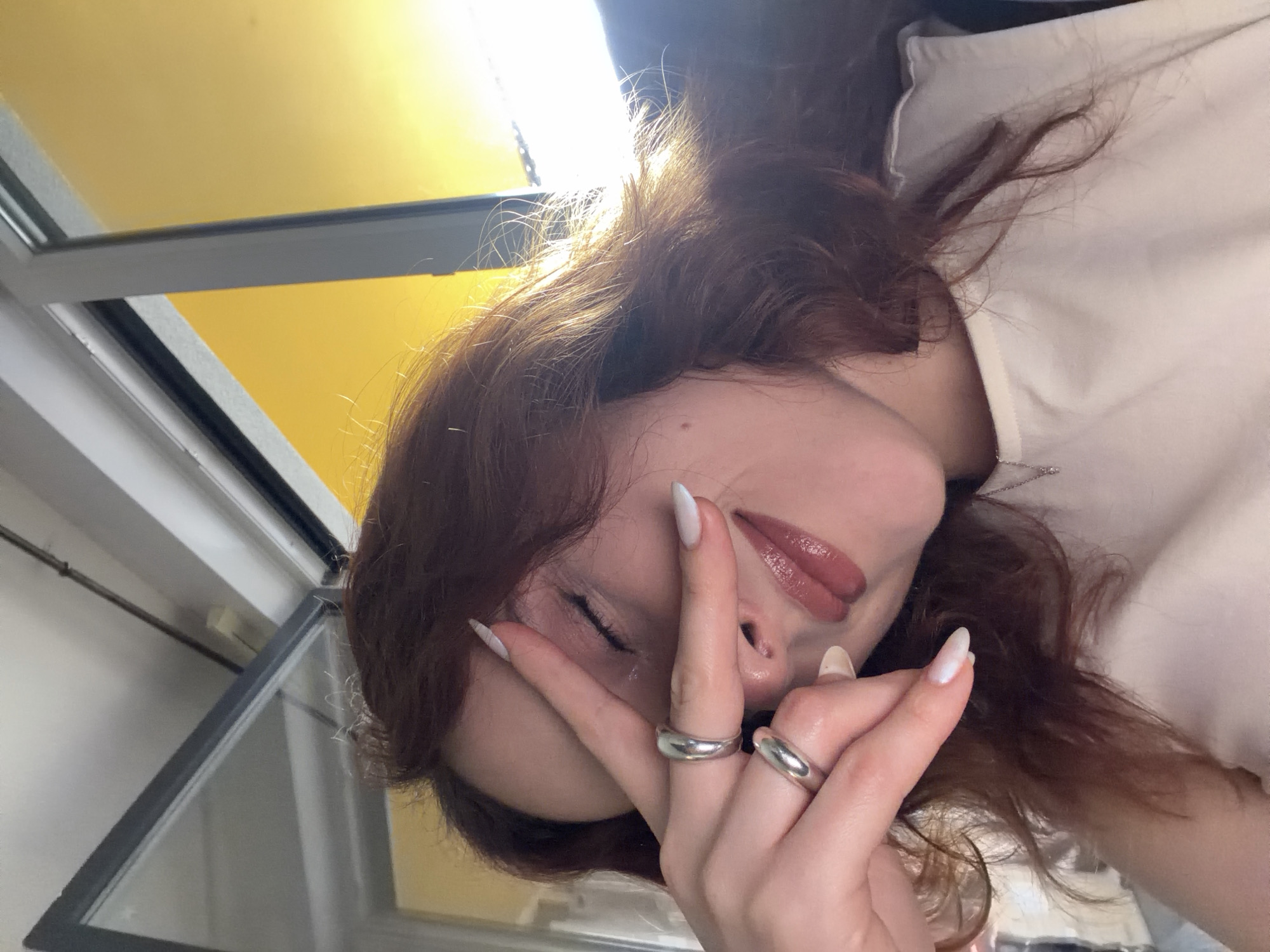 A woman with long, auburn hair and wearing a white shirt is positioned with her face near a window in Strasbourg, France. She is holding her hand up, displaying two silver rings on her fingers, while the other hand is visible in the background. Her lips are glossy and painted a mauve color. The window has a grey frame. The backdrop includes a yellow wall reflecting light. The overall angle of the photo is from slightly above the woman, looking downwards.