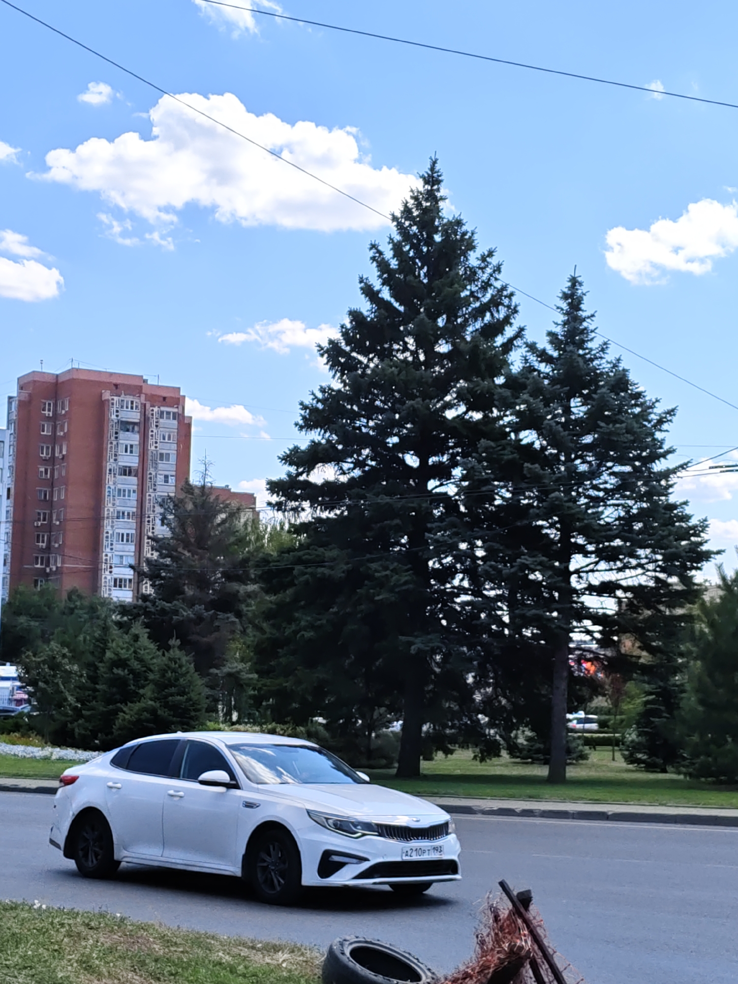 A white Kia Optima sedan is parked on a street in Rostov-on-Don, Russia. The car is facing to the right and is partially obscured by trees and foliage. In the background, a tall brick apartment building can be seen. The sky is blue with scattered white clouds. A traffic sign is visible in the foreground, as well as a discarded tire.