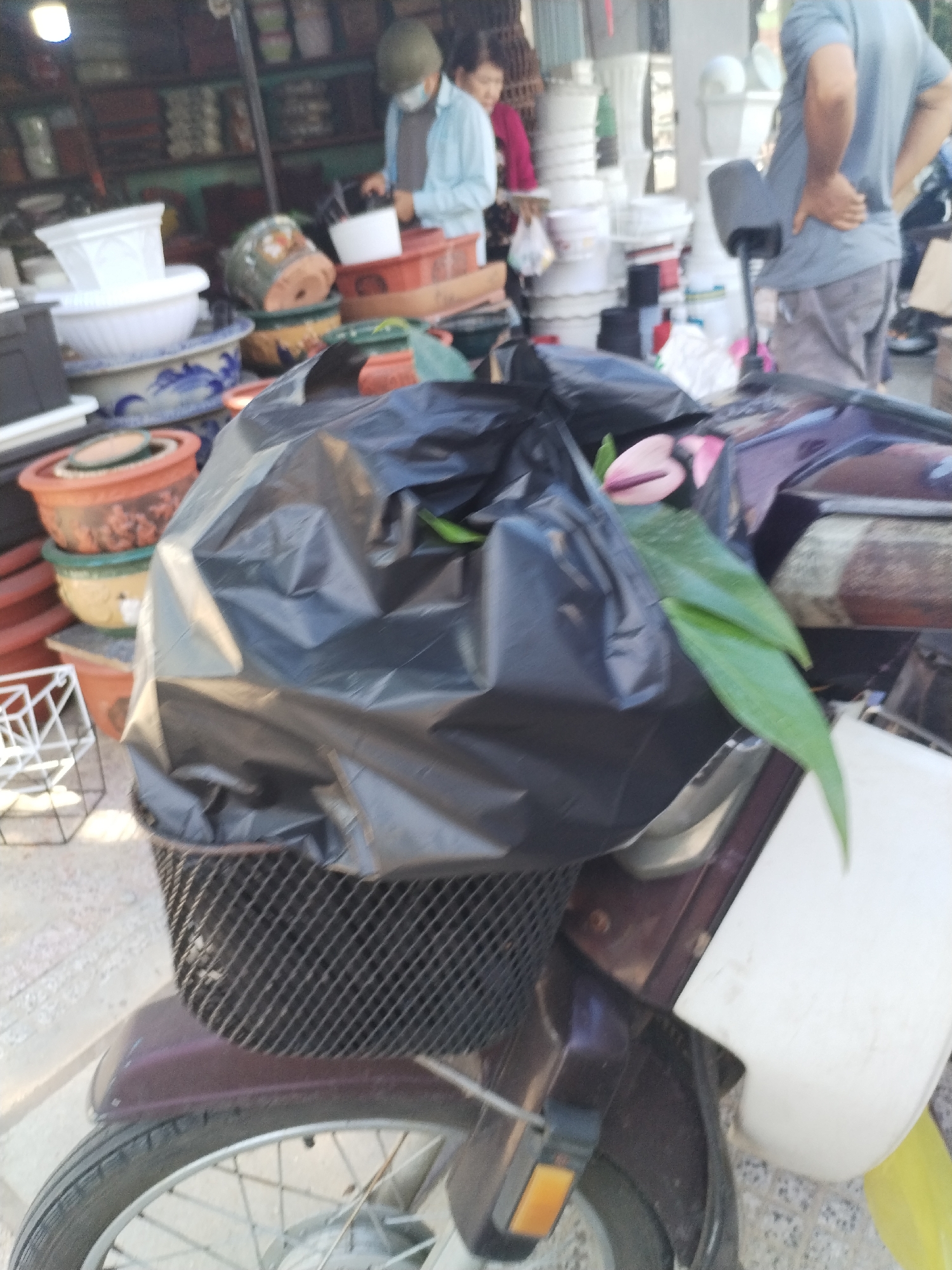 In a market stall in Bac Nha Trang Ward, Vietnam, a person wearing a light blue shirt and a green helmet is tending to goods. Another person, partially visible, stands behind them. In the foreground, a motorcycle with a black plastic bag filled with flowers and green leaves is parked. The bag is placed in the motorcycle's front basket. The stall is filled with various items, including stacked pots, buckets, and what appear to be garden supplies.