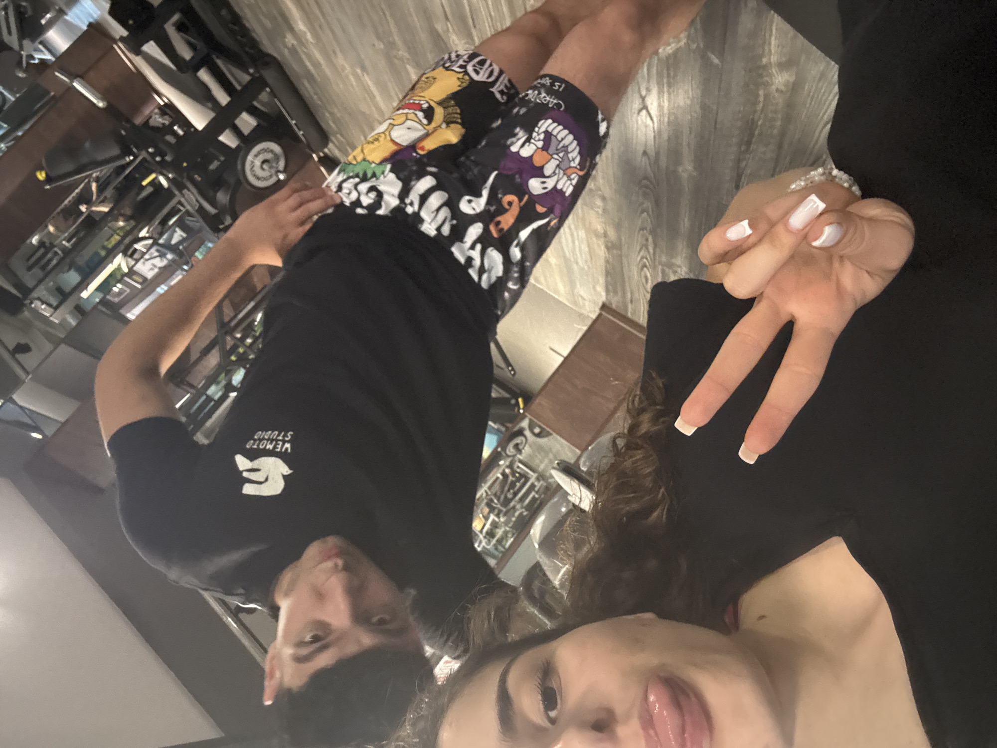 Two individuals, a female and a male, are taking a selfie in a gym. The female, positioned in the foreground on the left, is wearing a black t-shirt and has her tongue sticking out. Her right hand is raised, displaying a peace sign with her fingers. She has white manicured nails and a bracelet on her wrist. The male, on the right, is wearing a black t-shirt with a white logo and black shorts with cartoon prints, including characters resembling those from "The Simpsons." He is looking at the camera with a serious expression. The background shows gym equipment, suggesting the location is a fitness center. The lighting is typical of an indoor gym environment. The image does not provide any specific information about the city of Shumen, Bulgaria.