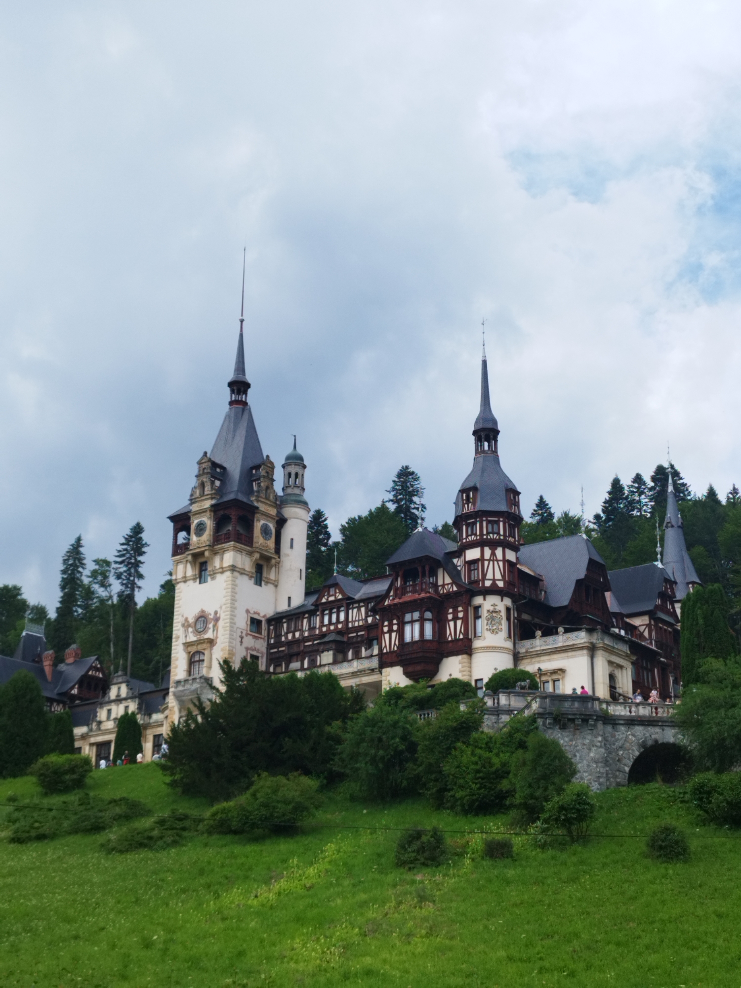 This image captures a view of Peles Castle, a historic landmark in Sinaia, Romania, situated amidst lush green rolling hills and dense forests. The castle, with its striking neo-Renaissance architecture, is prominently featured with its numerous towers and spires reaching towards a cloudy sky. The main tower, a focal point of the castle, displays a clock face. The castle's facade exhibits a mix of cream-colored walls and dark timber framing, characteristic of its architectural style.
In the foreground, a verdant lawn slopes gently upwards towards the castle. Scattered across the lawn are various bushes and trees, some of which are quite dense and tall. In the middle ground, a stone bridge arches over a lower section of the terrain, leading to a pathway that runs alongside the castle. A few individuals, appearing as small figures, are visible on the bridge and on a balcony of the castle, suggesting visitor activity. The background is dominated by a dense forest of pine trees, forming a dark green backdrop against the overcast sky. The overall scene conveys a sense of historical grandeur and natural beauty.