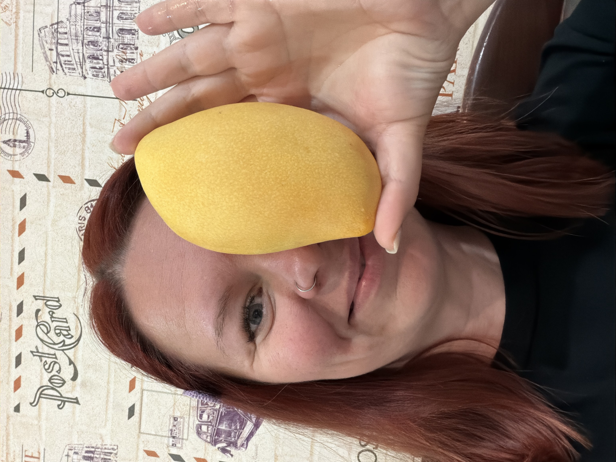 A woman with red hair holds a yellow mango in front of her face. She is wearing a nose ring and a black top. The background is a patterned surface with postcard designs. The location is Chaikovsky, Russia.