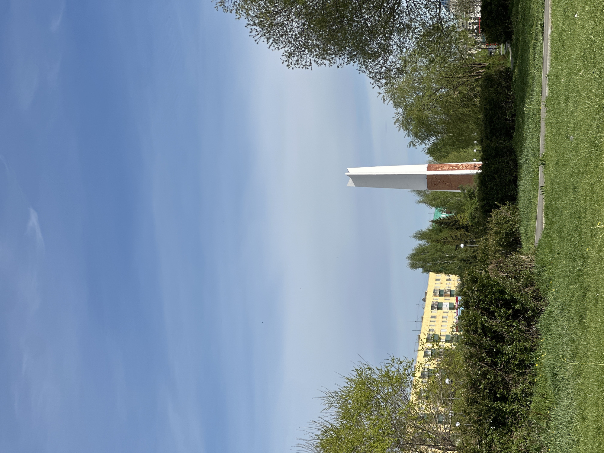 The image shows a tall white obelisk with a brown relief, possibly a war memorial, framed by trees and bushes. A yellow building with multiple windows is visible behind the foliage. The foreground is dominated by green grass, and the sky above is a clear blue with some cloud streaks. The location is in a city with urban elements.