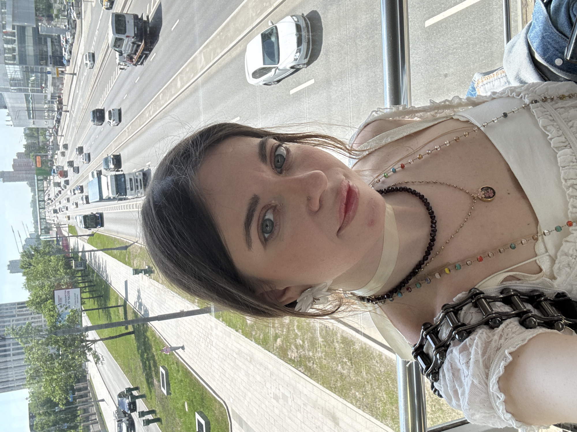 A selfie of a woman with green eyes and dark hair is oriented at an angle with a cityscape visible in the background. The scene appears to be taken from a high vantage point, possibly inside a building looking out over a multi-lane highway with cars and trucks. On the left side of the frame, there is a pedestrian path with trees and buildings. The woman is wearing a white top with lace detailing and several necklaces, including a beaded choker and a chain-link bracelet. The location is Moscow, Russia, indicated by signage in Russian in the background.
