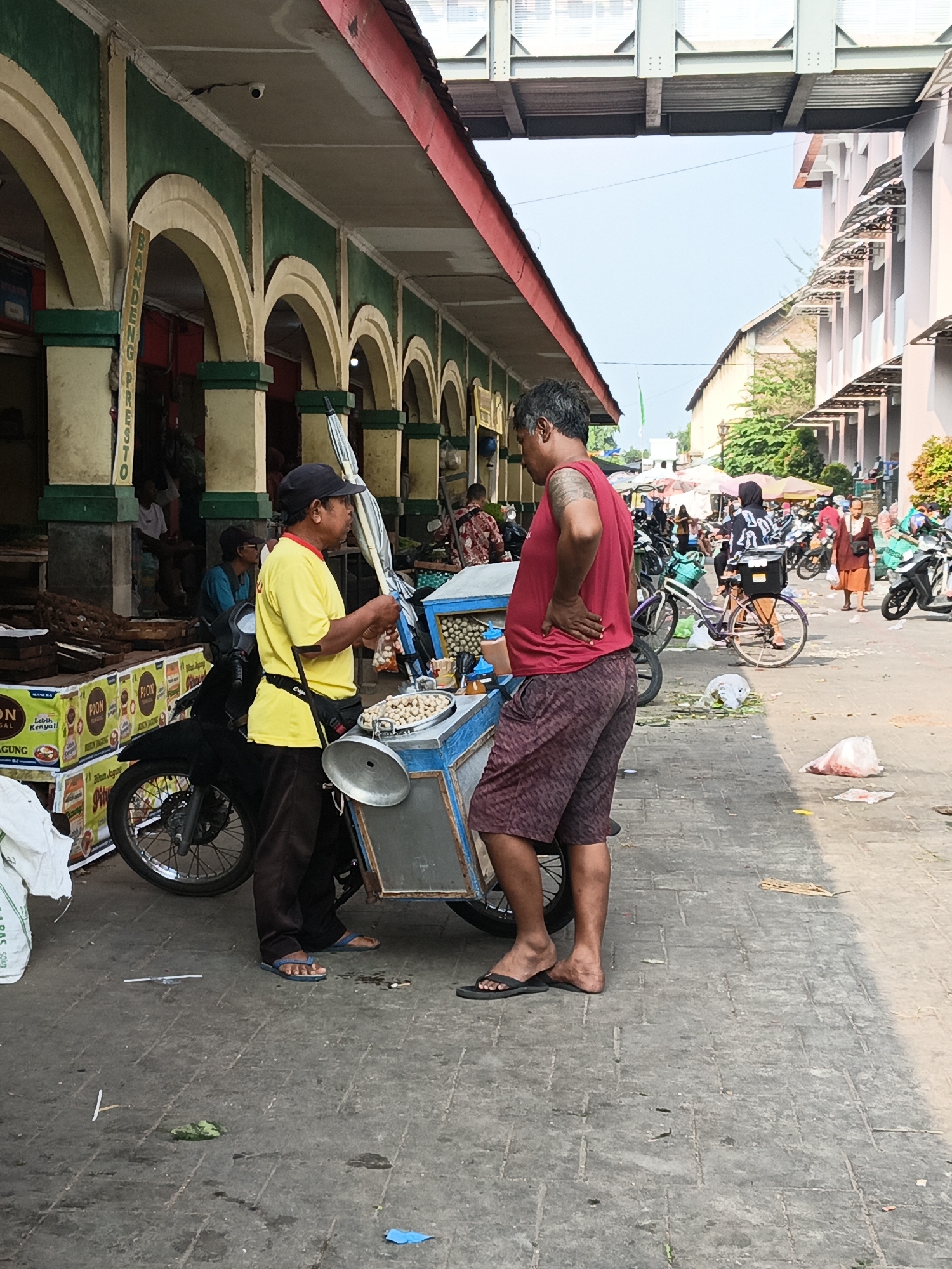 The image depicts a busy outdoor market scene in Klaten, Indonesia, likely along a street or thoroughfare. In the foreground, two men are engaged in conversation. One man, wearing a yellow t-shirt and dark pants with blue flip-flops, stands by a food cart. He holds a bag of items and appears to be interacting with the other man. The second man, wearing a red sleeveless shirt and patterned shorts with black flip-flops, stands with his hand on his hip, facing the man in yellow. His left arm has a tattoo.
The food cart is blue and silver, with a large metal bowl on top containing a pile of small, round, light-colored food items, possibly snacks or ingredients. A black motorcycle is parked next to the cart. Behind the cart, stacks of yellow and red boxes with the "PION" logo are visible, suggesting a commercial establishment.
The environment features an arched walkway with green and yellow columns, indicative of traditional market architecture. Various other market-goers, bicycles, and motorcycles are present in the background, suggesting a lively and bustling atmosphere. The ground is paved with interlocking bricks. The sky is clear and bright, indicating daylight. The overall impression is of a typical Indonesian street market.