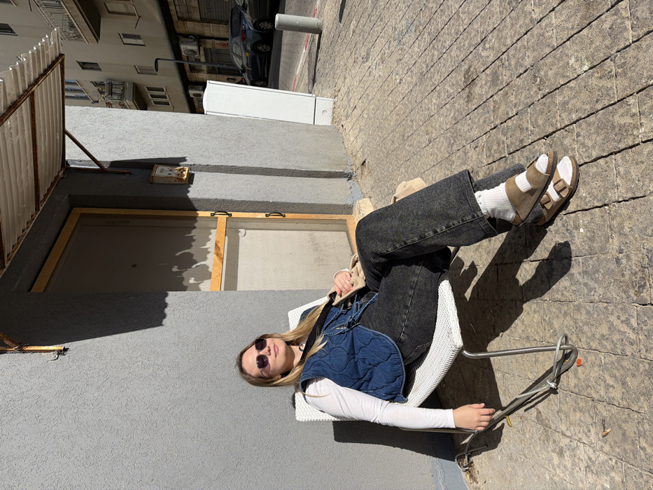 A female individual is seated on a white metal-framed chair on a paved sidewalk in Tel-Aviv, Israel, facing to the viewer's right and slightly upward towards direct sunlight. She is wearing dark sunglasses, a white long-sleeved top, a blue quilted denim vest, dark grey wash jeans, white socks, and brown open-toed sandals. A light beige shoulder bag with a black strap is visible across her chest. Her long, light-colored hair is down.
Behind her, a grey-textured building facade features a brown-framed door, possibly a screen door, partially visible. Above the door, a rusted corrugated metal awning is supported by rusted brackets. To the right of the door, a small, weathered yellow mailbox or utility box is mounted on the wall. The ground beneath the chair and extending into the foreground is paved with irregularly shaped grey and brown stones. Grey cables are visible on the ground near the chair's front leg.
In the background, a street runs parallel to the building, occupied by parked cars, including a dark blue vehicle. Several grey cylindrical bollards line the edge of the sidewalk. Further back, a multi-story residential building with numerous windows, balconies, and external air conditioning units is visible under bright daylight, casting distinct shadows from the building and the seated person. A white rectangular utility box stands on the sidewalk to the right.
