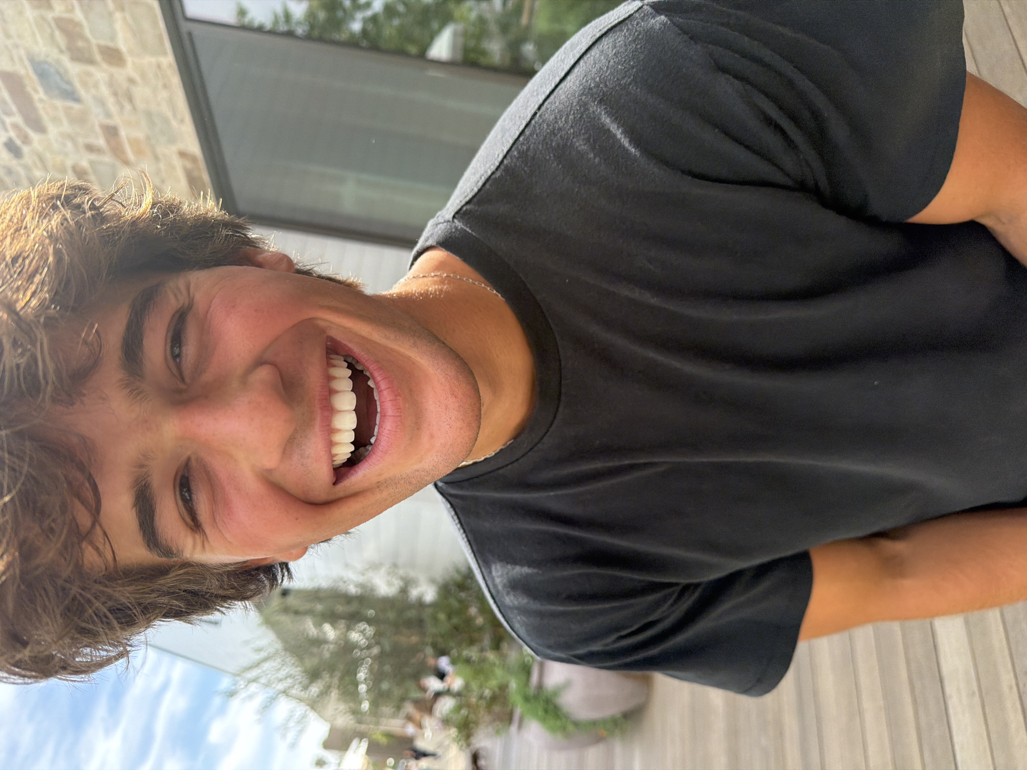 A medium close-up, slightly angled view depicts a smiling young man, possibly in his late teens or early twenties, wearing a dark t-shirt and a silver necklace. The setting appears to be outdoors during daylight, evidenced by the natural light illuminating the subject and the visible sky.
The background features a blurred view of trees, a building with a large window, and a partial view of stone exterior. Elements like potted plants suggest a patio or garden. Given the description, this image likely originates from Malibu, California, United States.
