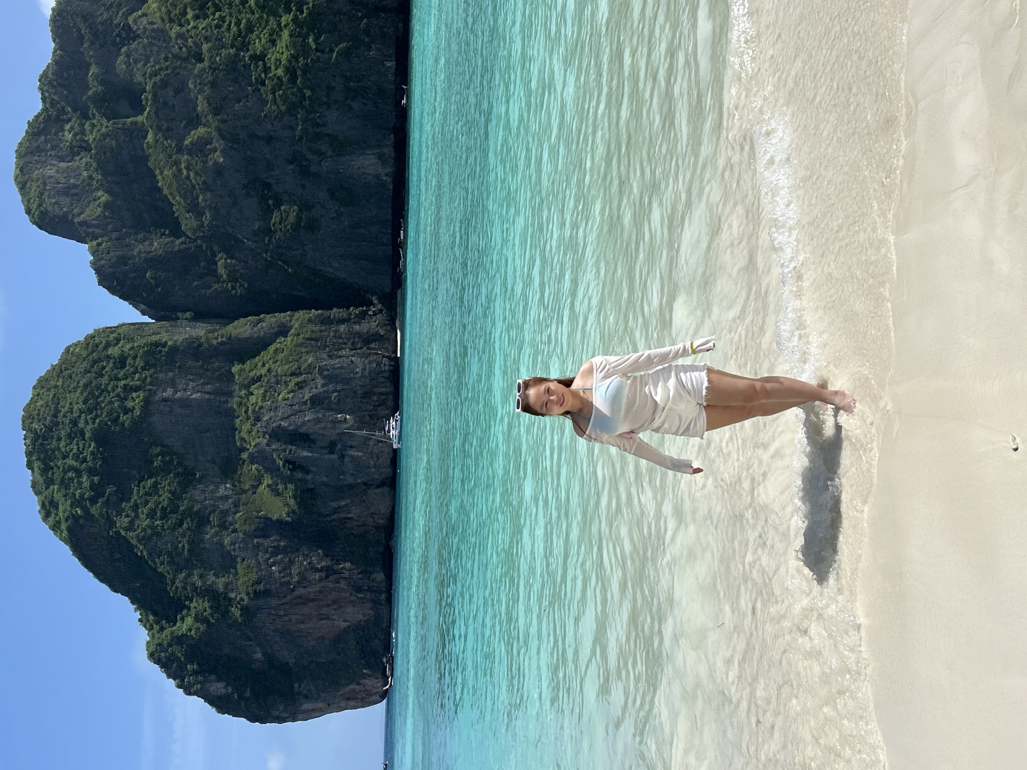 A young woman stands in shallow, turquoise water on a sandy beach. She is facing the camera and smiling, wearing a white off-the-shoulder top and white shorts. Her hair is dark and pulled back, and she has sunglasses on her head. Behind her, the ocean stretches out to large, jungle-covered limestone karsts rising from the water. A sailboat and smaller boats are visible in the distance. The sky is clear and blue. The scene appears to be Maya Bay, located in Krabi Province, Thailand, a popular tourist destination accessible from Ao Nang.