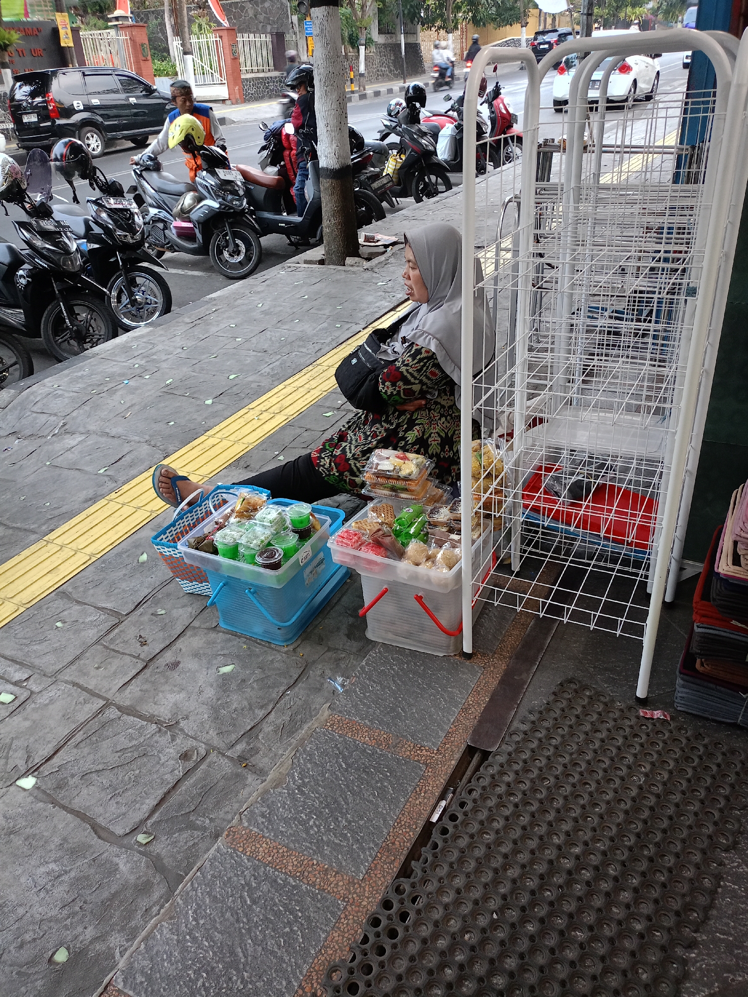 A woman wearing a grey hijab and a patterned dress is sitting on a sidewalk in Batu, Indonesia, beside a white wire display rack. She is partially facing away from the camera, with her right leg extended. In front of her are two blue plastic baskets and a clear plastic container filled with packaged food items, including drinks and snacks. The sidewalk has a yellow tactile paving strip for the visually impaired. Behind the woman, several motorcycles are parked along the street, and a black SUV is visible. The environment appears to be an urban setting with buildings and trees in the background, suggesting a commercial or roadside area.