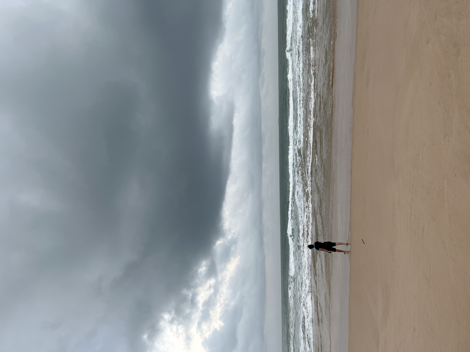 A lone individual stands at the edge of the water on a beach in Habaraduwa, Sri Lanka, facing the ocean under a dramatic, overcast sky. The person, seen from behind, is wearing dark shorts and a dark t-shirt with a blue collar. The sea is choppy with white-capped waves breaking onto the sandy shore. The sky is filled with heavy, dark gray clouds, suggesting an impending storm or recent rainfall. The sand is a light brown, appearing wet near the water's edge. The overall atmosphere is subdued and somewhat somber due to the weather conditions.